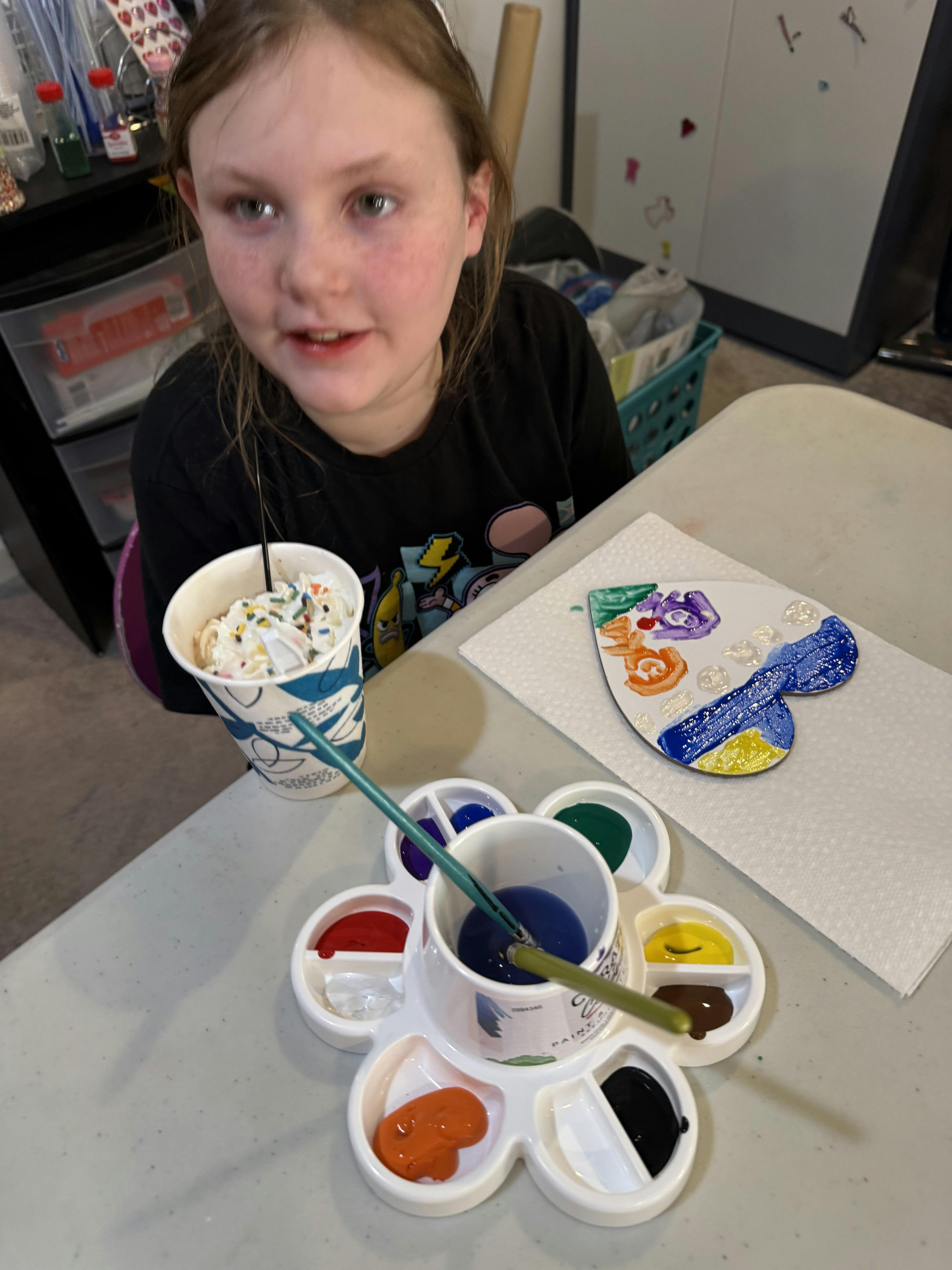 Child painting a heart-shaped object, holding a drink with whipped cream