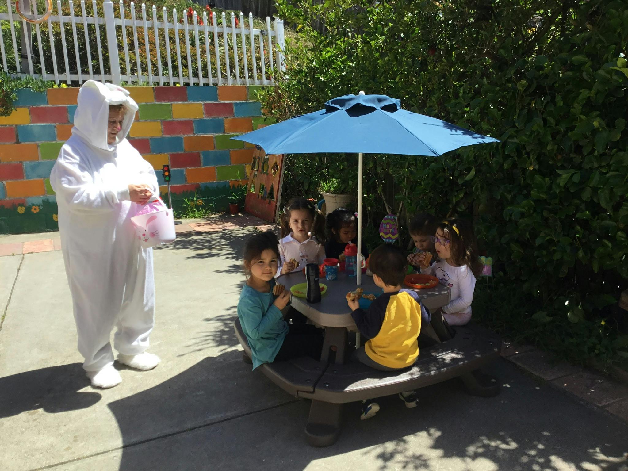Person in white costume holding a bag, children sitting at a picnic table under an umbrella