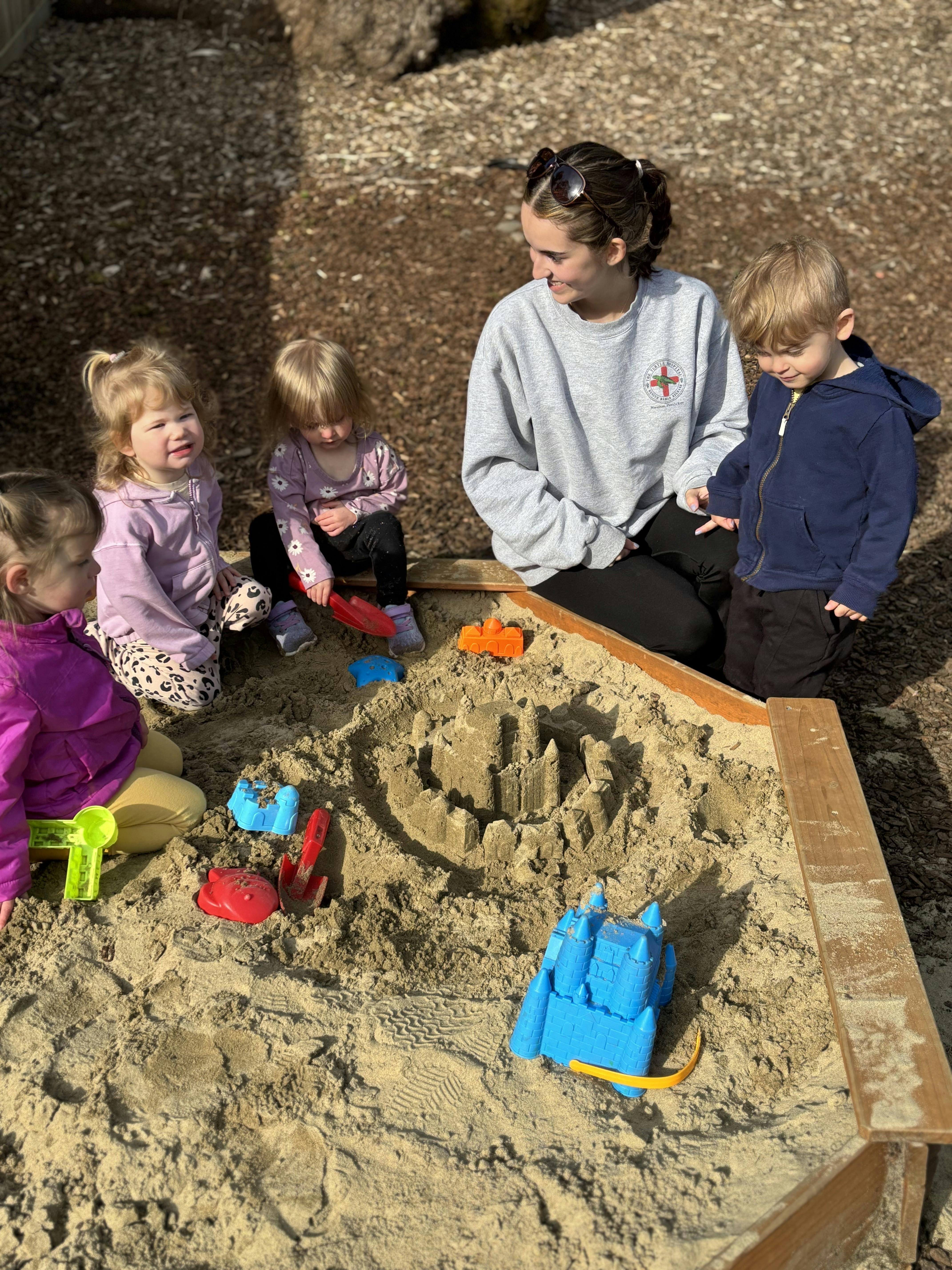A person and four children playing in a sandbox with a sandcastle