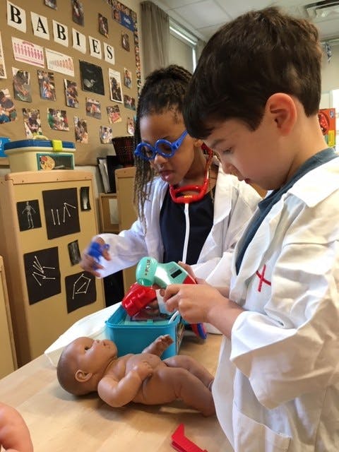 Two children in lab coats playing with a baby doll and toy medical tools