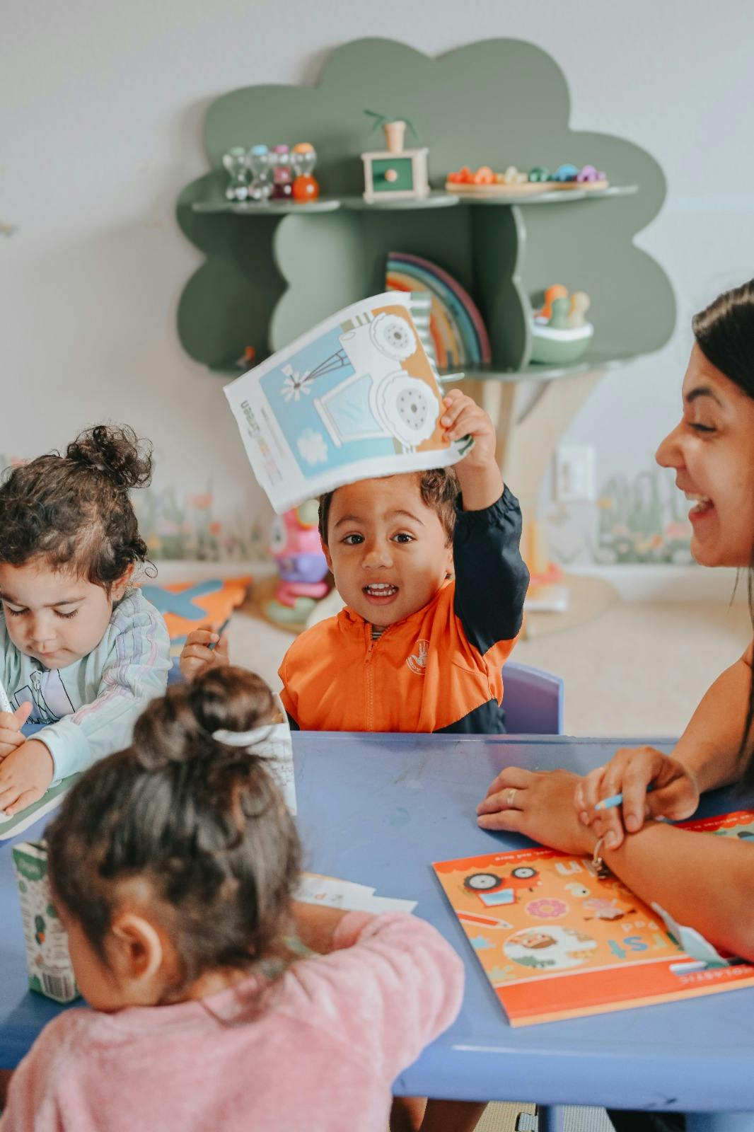 Child holding a book over their head in a classroom with other children and an adult
