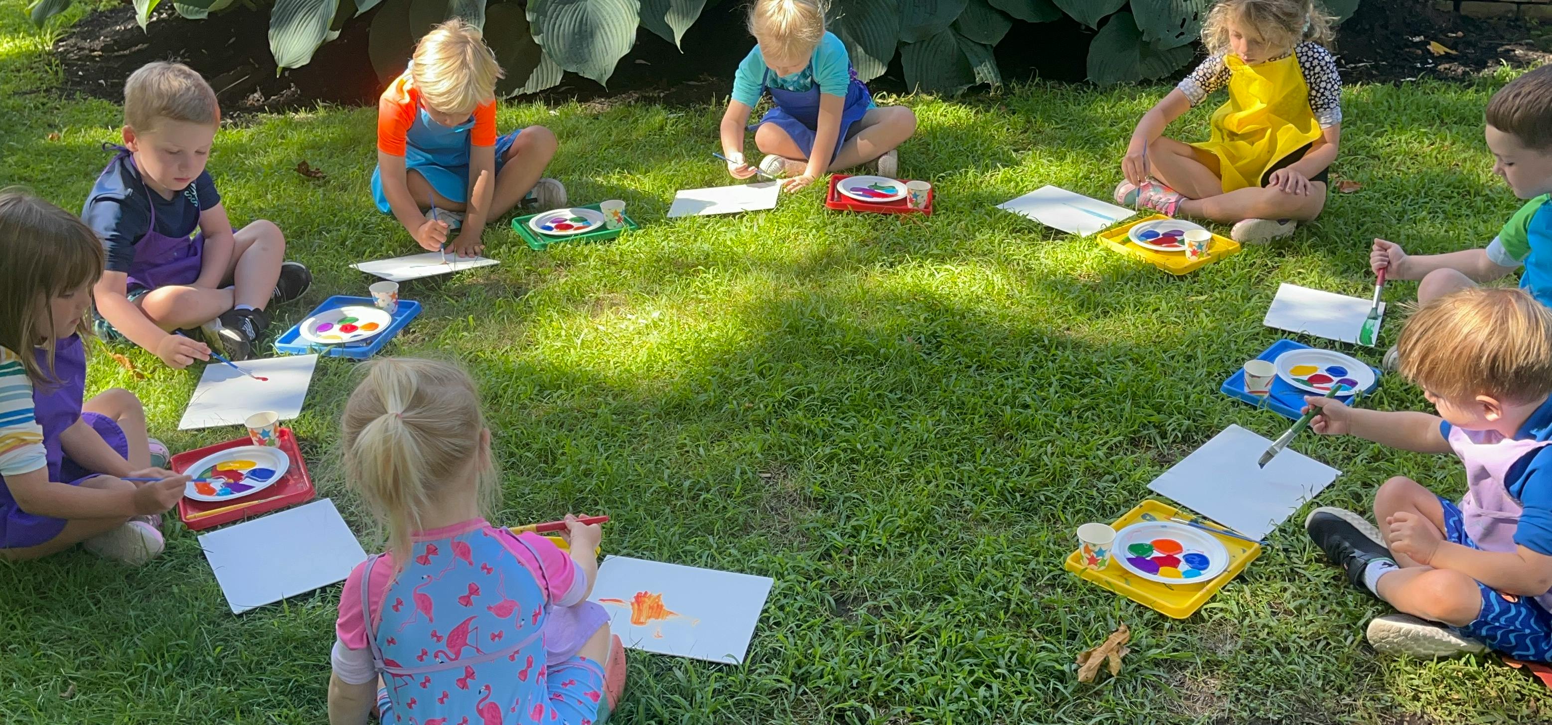 Children sitting in a circle on grass painting on paper with brushes