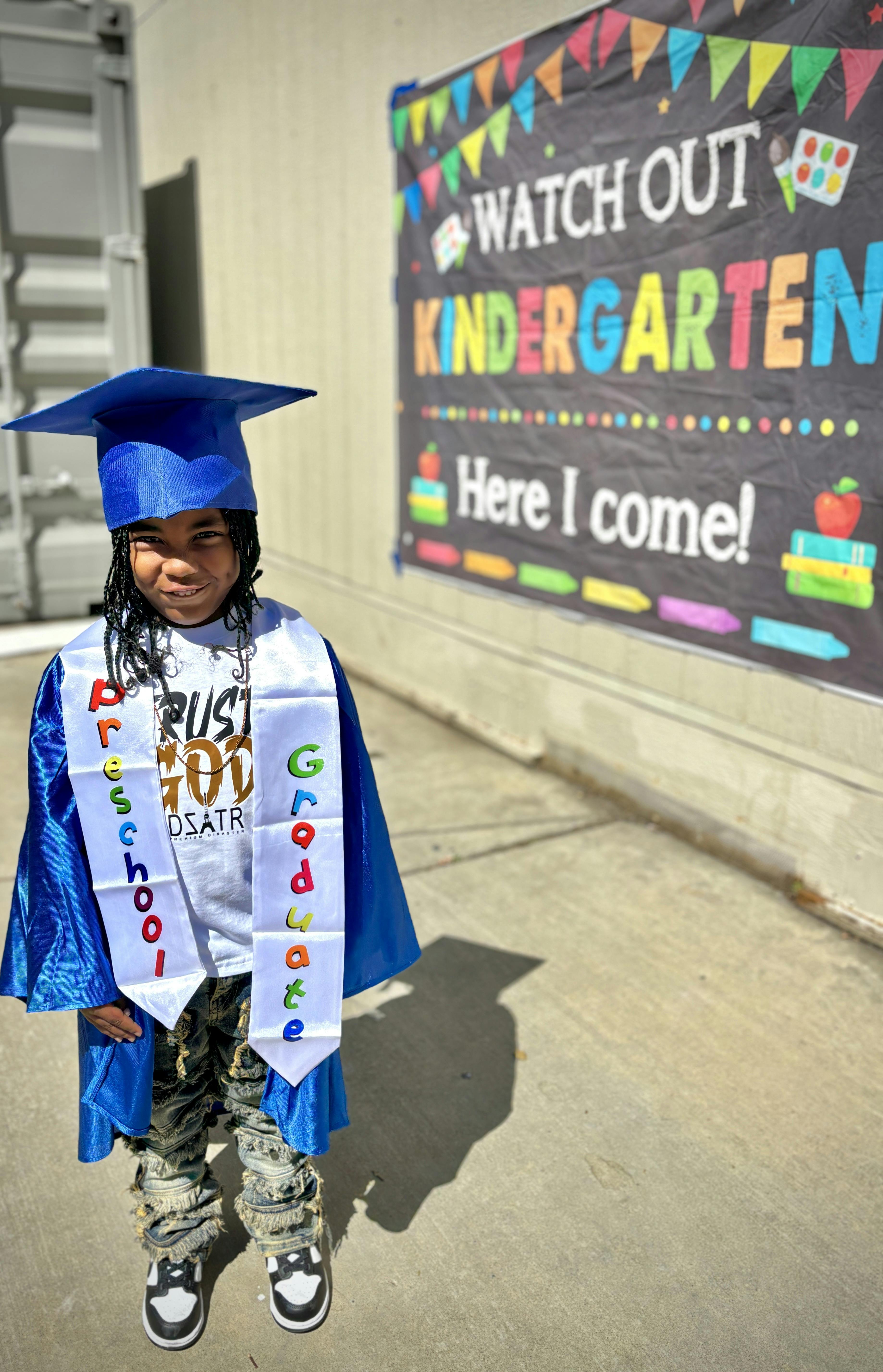 Child in blue graduation cap and gown with a "Preschool Graduate" sash