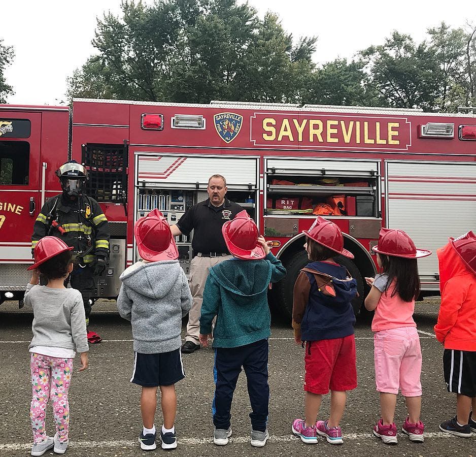 Children in red hats looking at firefighters and a fire truck with "SAYREVILLE" on it