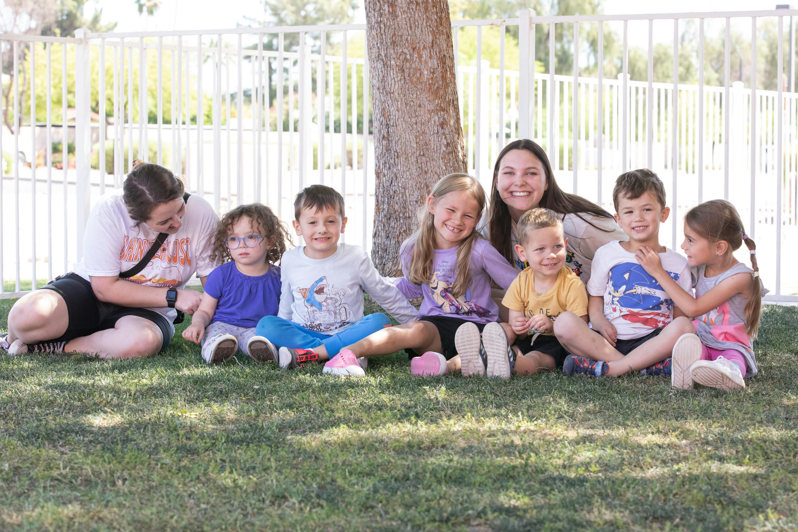 Group of two people and six children sitting on grass in front of a tree
