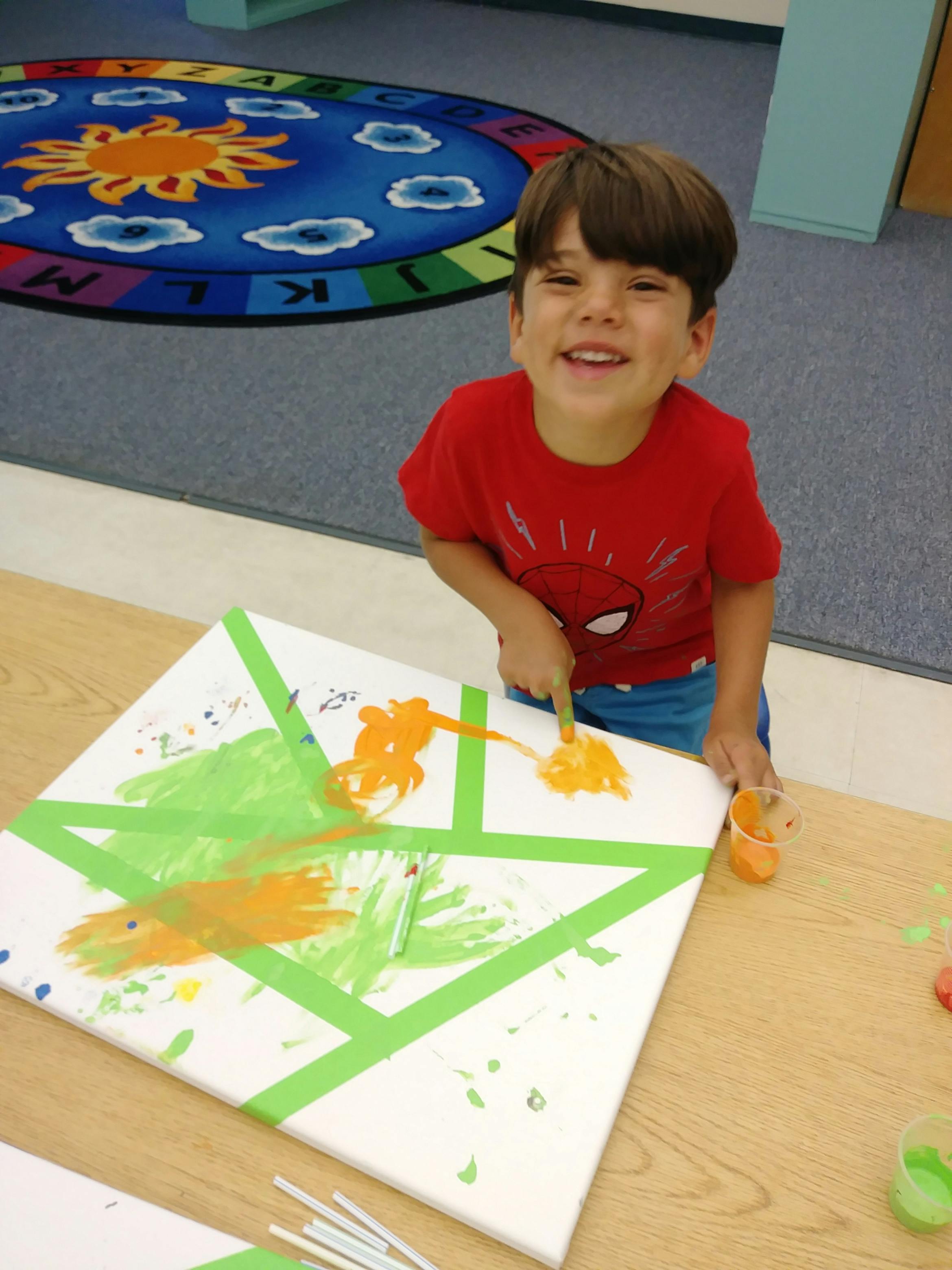 Child painting on a canvas with green tape, using fingers to apply orange paint