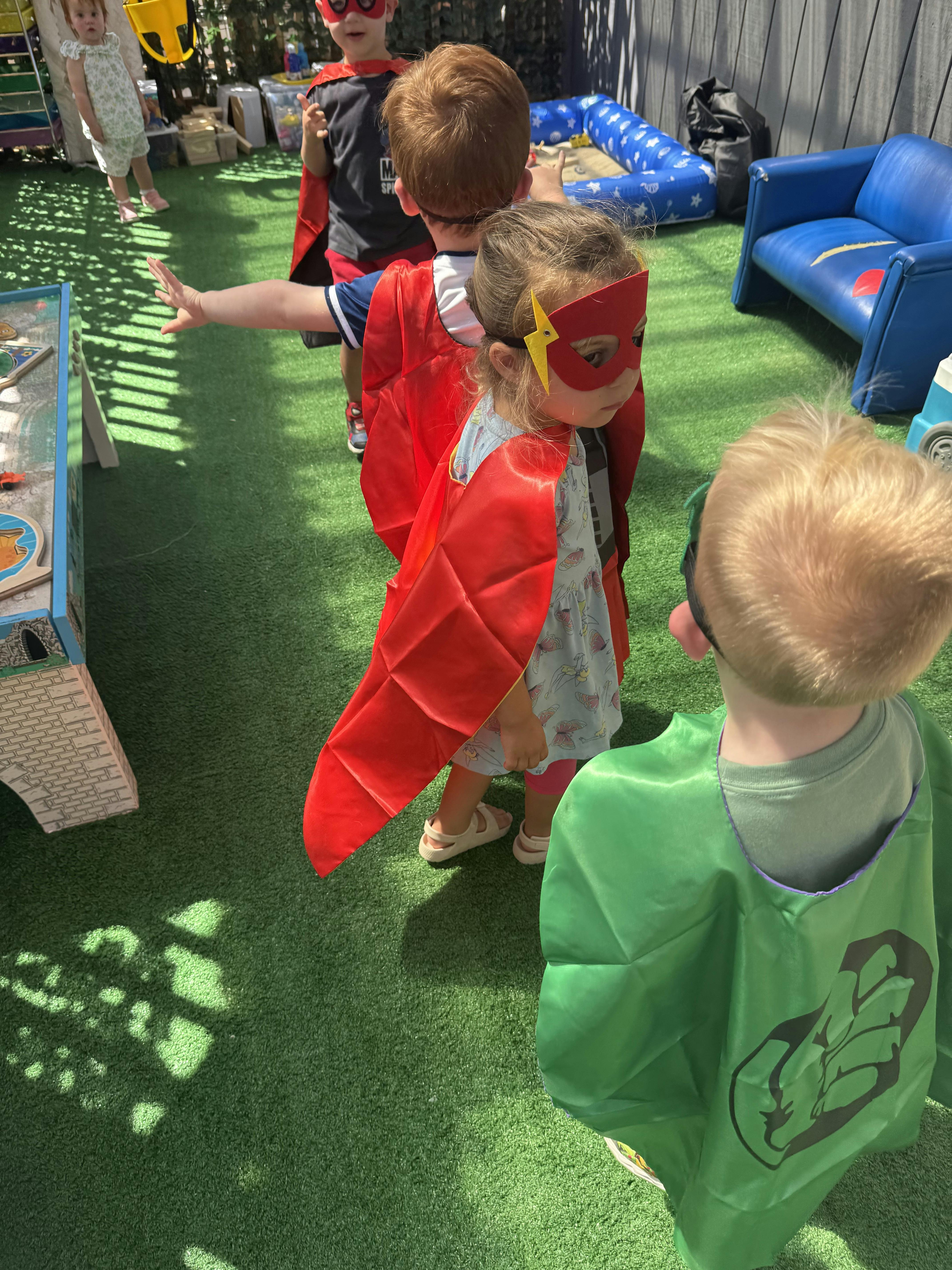 Children wearing superhero costumes and capes standing on artificial grass