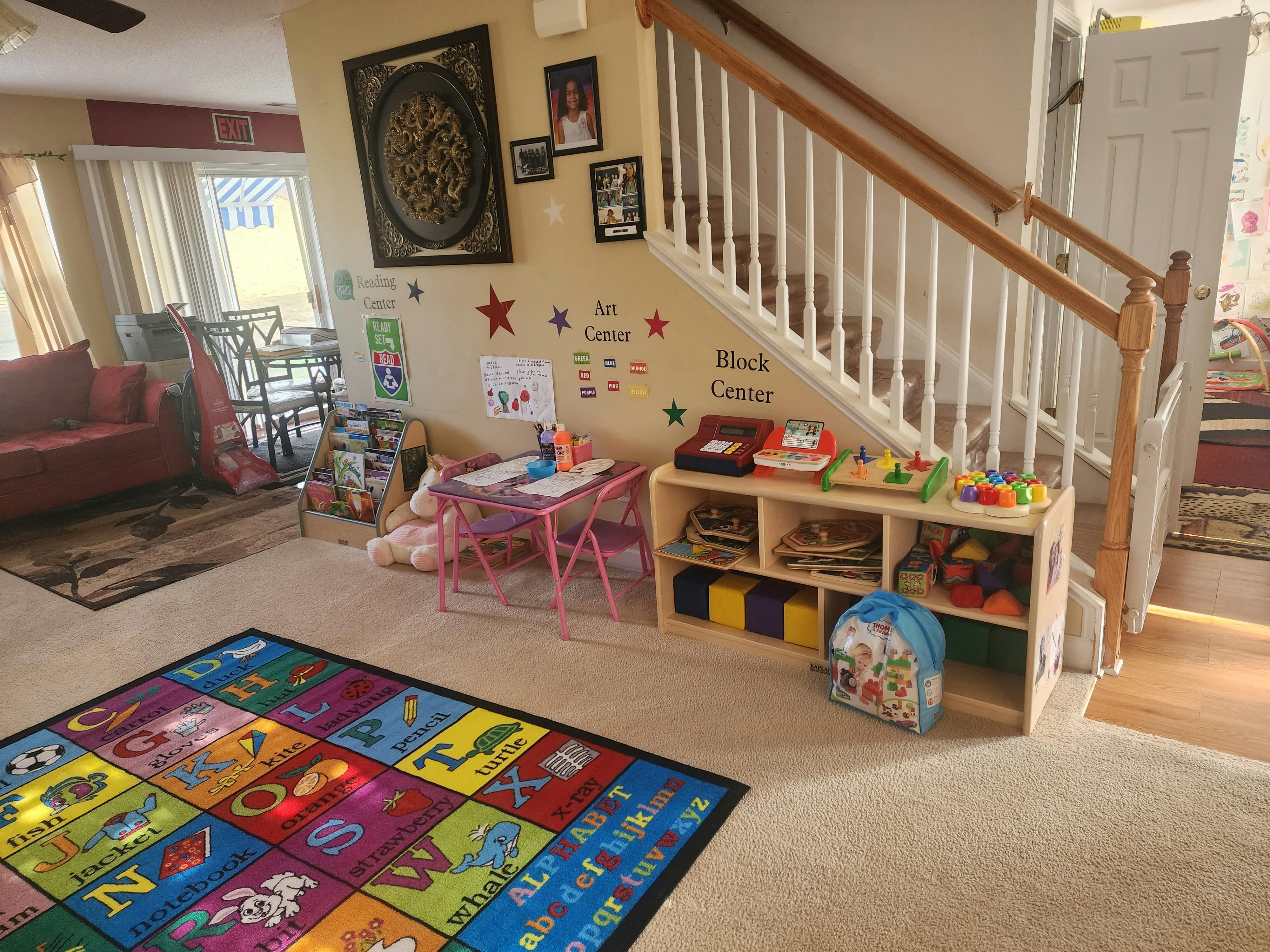 Playroom with alphabet rug, small table, and toy shelf next to a staircase