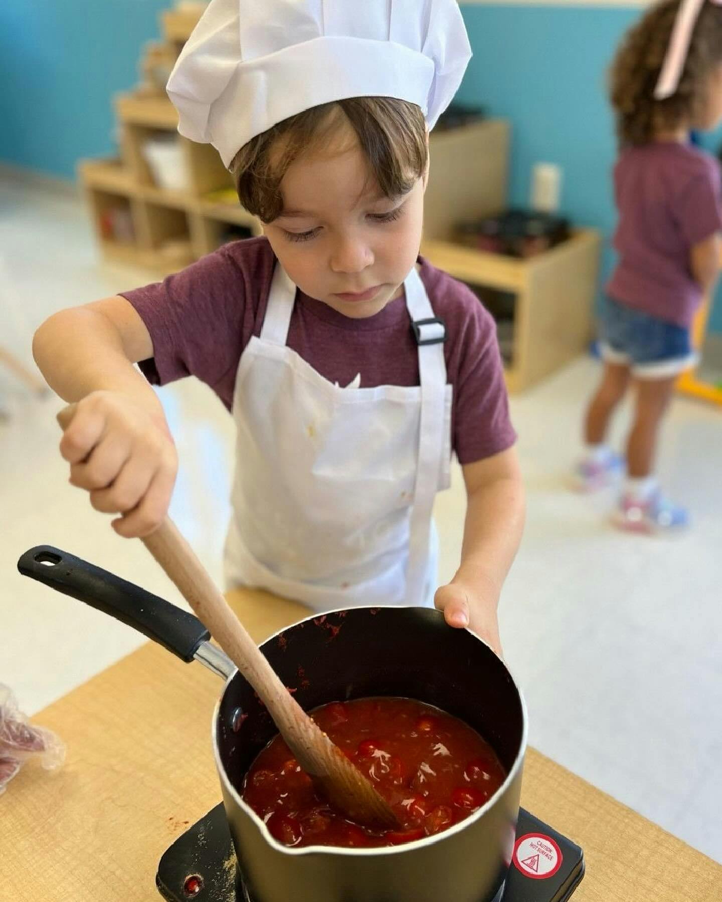 Child in chef hat and apron stirring red sauce in a pot