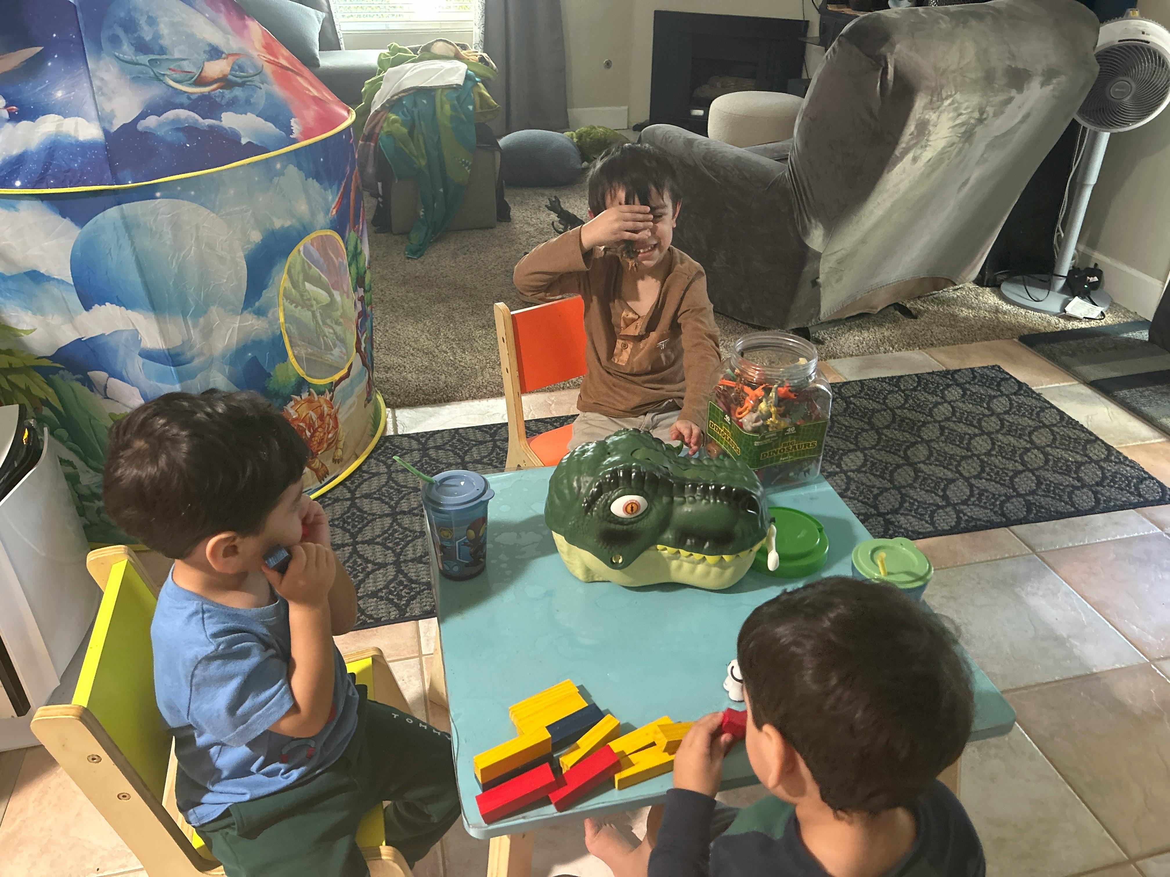 Three children playing at a small table with toys, one covering eyes