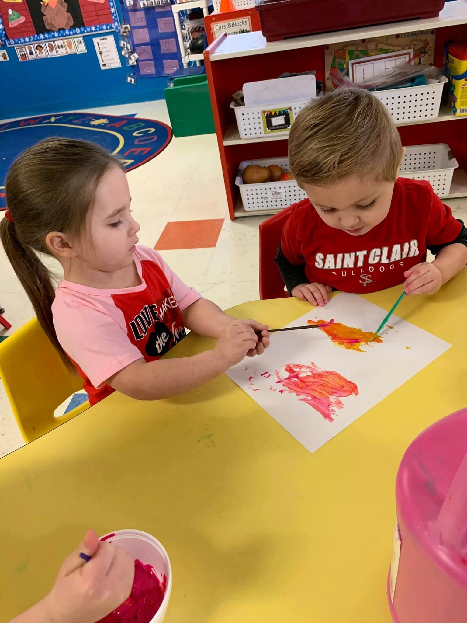 Two children painting at a yellow table in a classroom