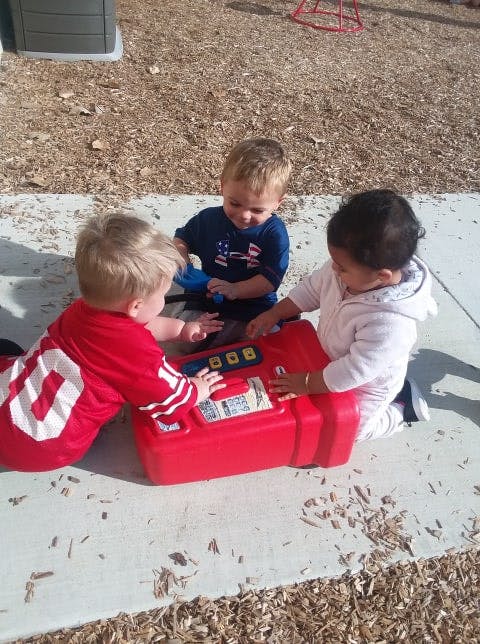Three children playing with a red toy cash register outdoors