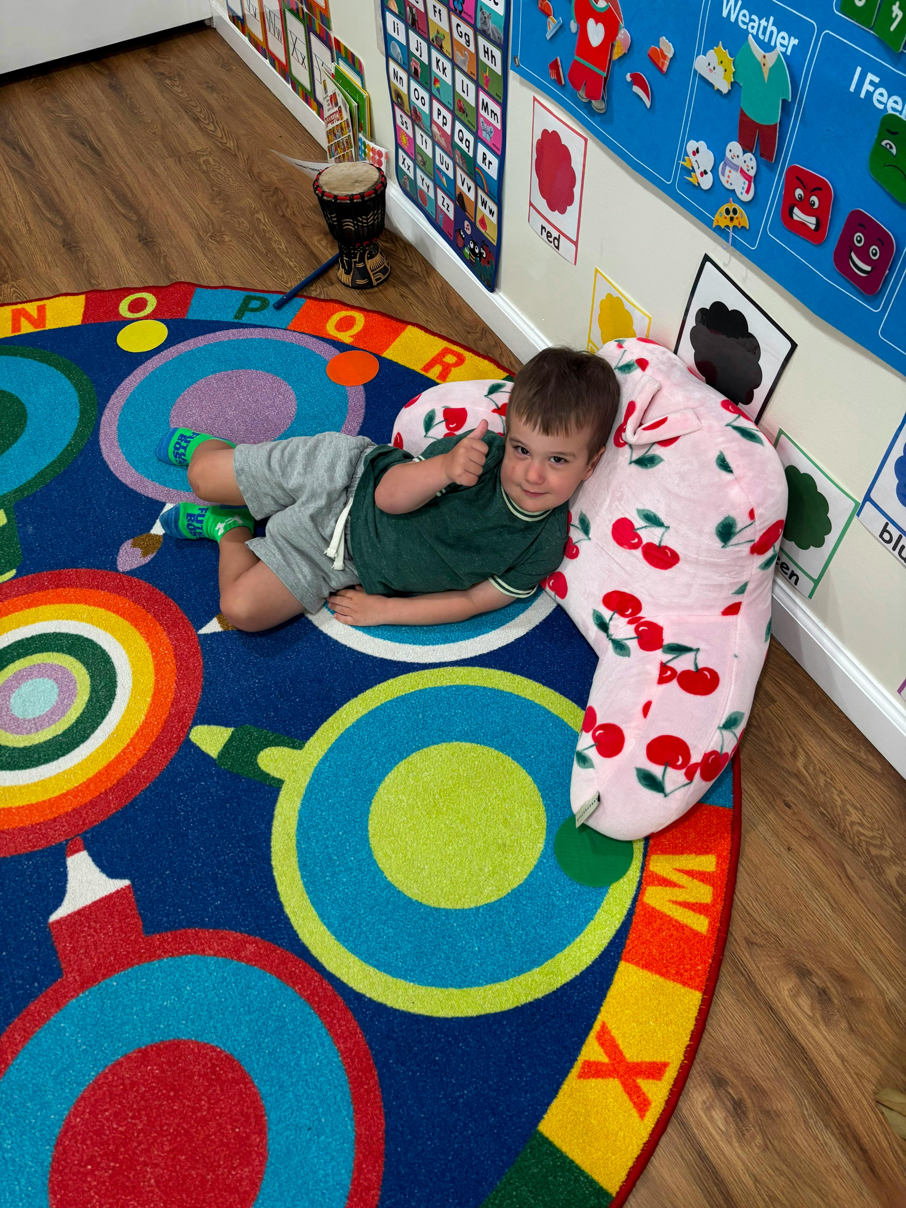 Child lying on a classroom rug against a cherry-patterned pillow giving a thumbs up