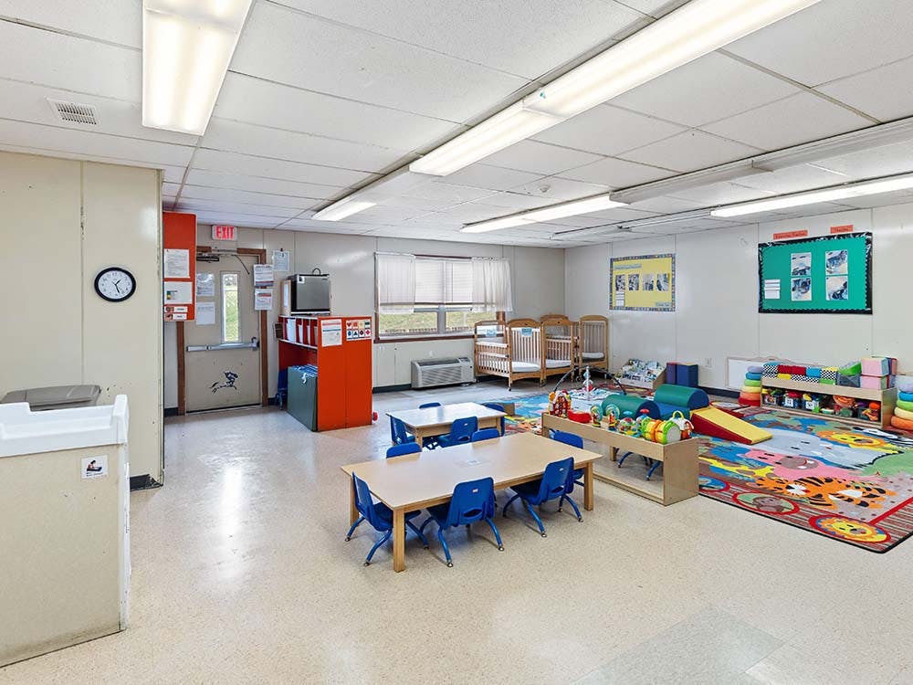 Classroom with small tables and chairs, play mats, and several cribs