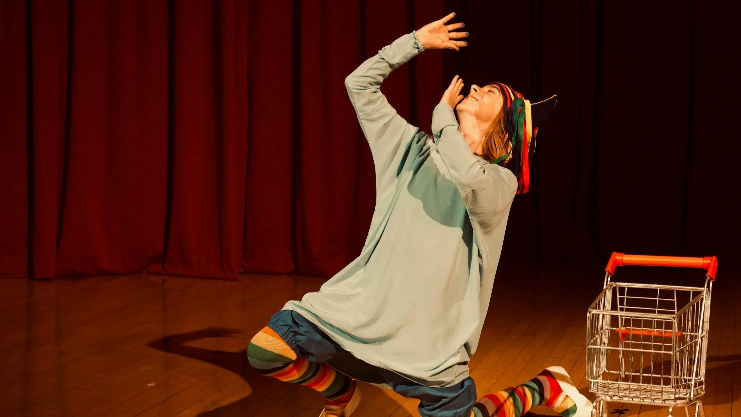Person in colorful costume and hat on stage with a toy shopping cart