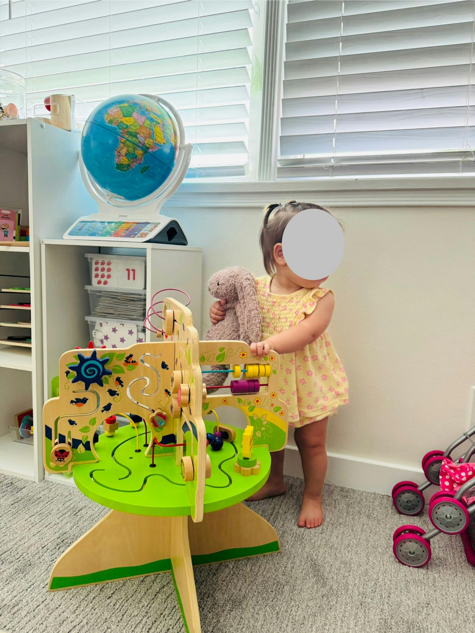 Child holding a stuffed animal and standing next to a wooden activity table