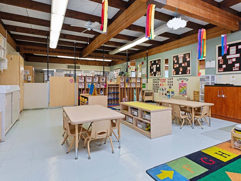 Empty classroom with small tables and chairs, shelves with toys, and alphabet art