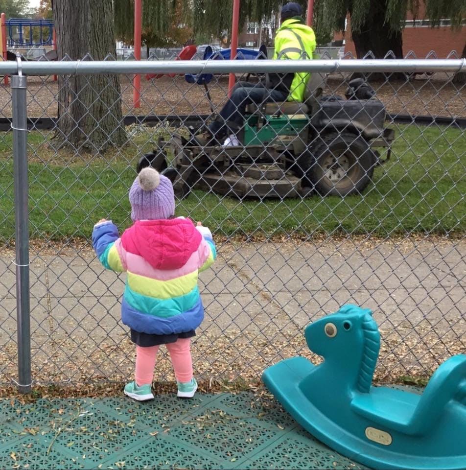 Child in colorful coat looking through a fence at a person on a lawnmower