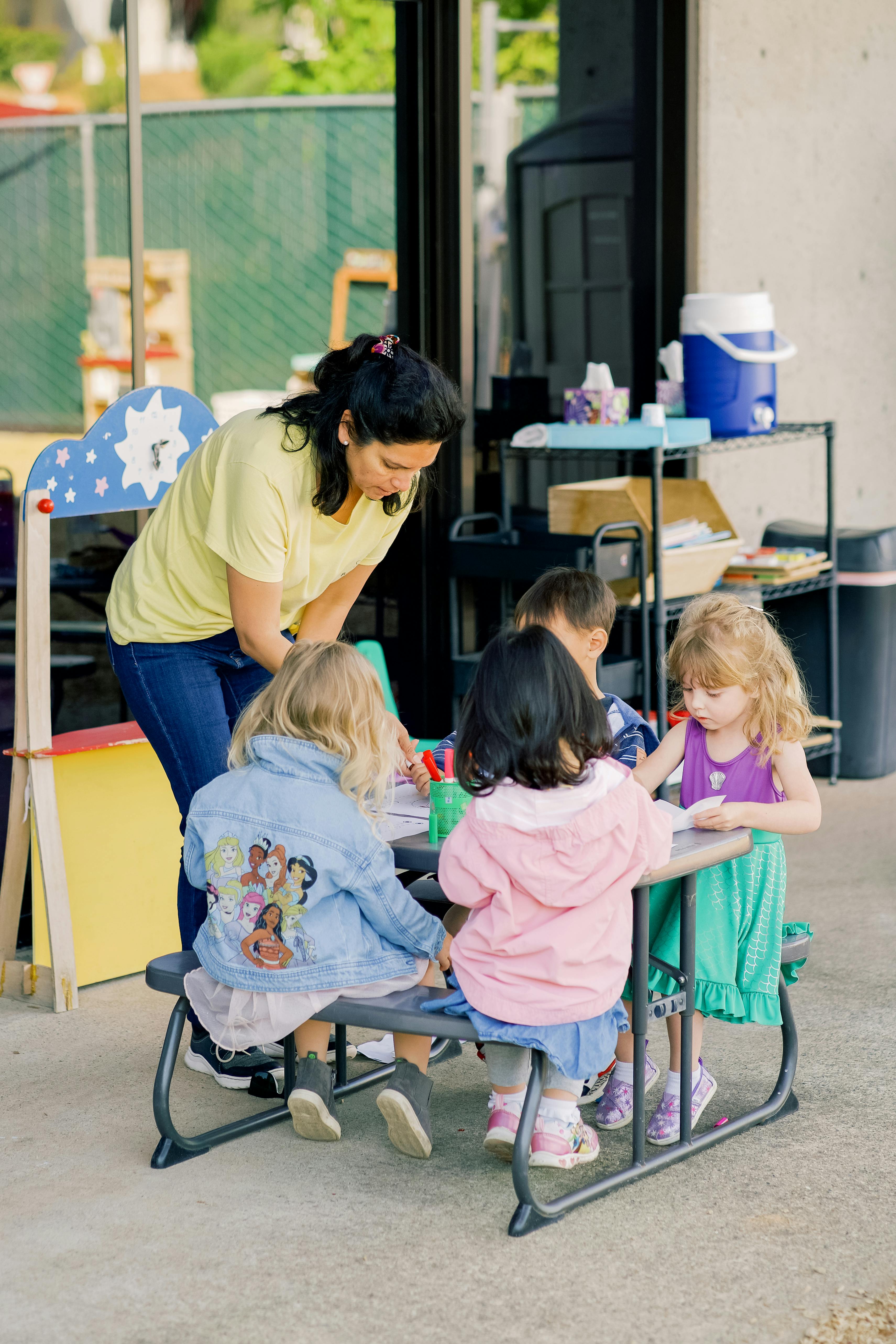 Adult supervising children drawing at a small table outdoors