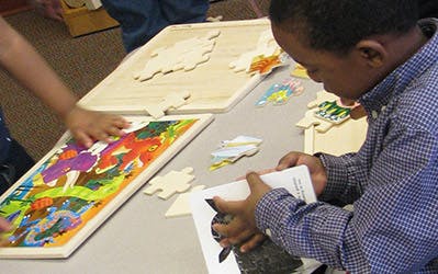 Child looking at a book while another child works on a puzzle