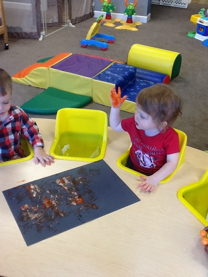 Child with orange paint on hand at a table with another child and artwork