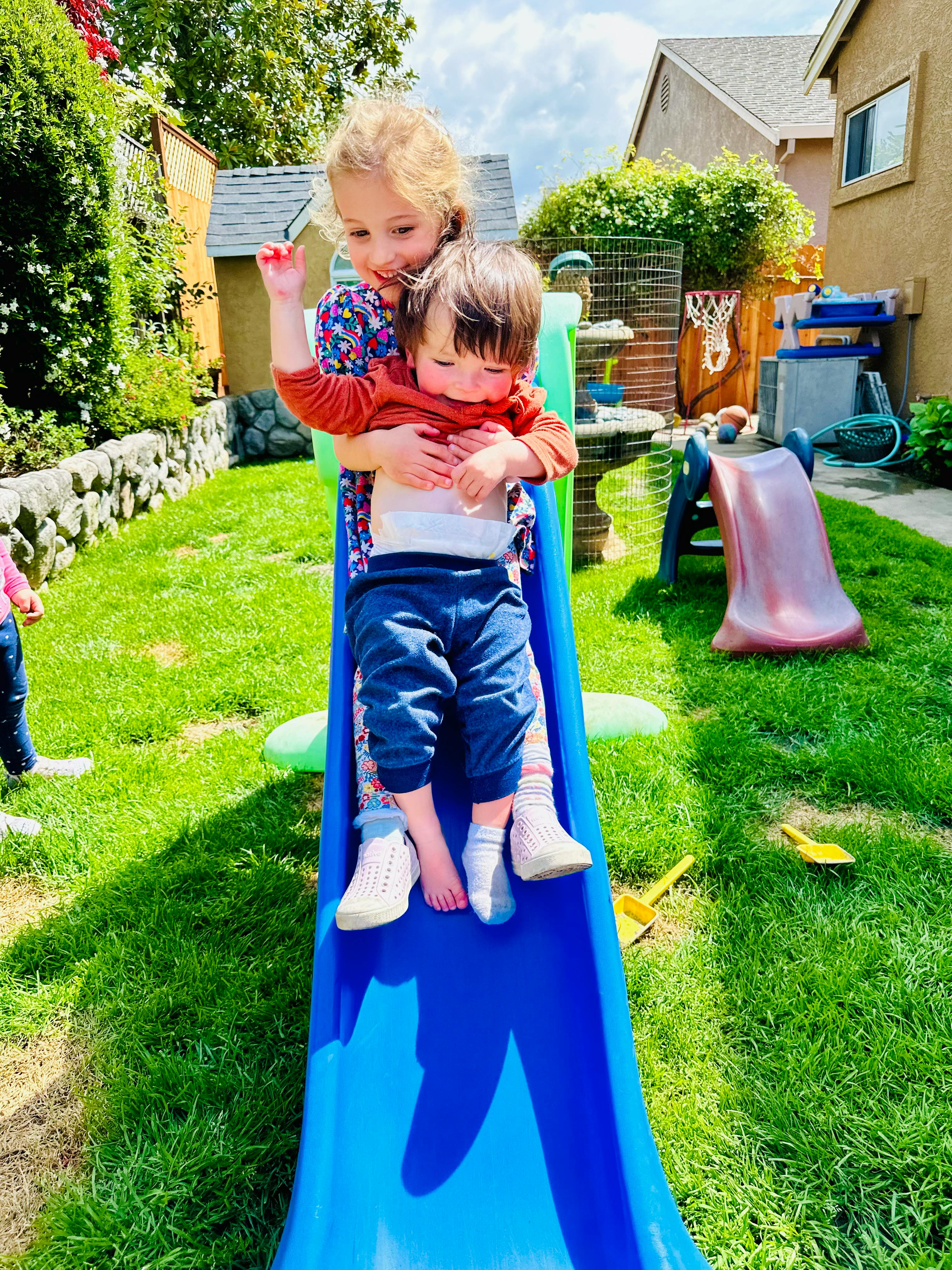 Child holding a smaller child on a blue slide in a backyard