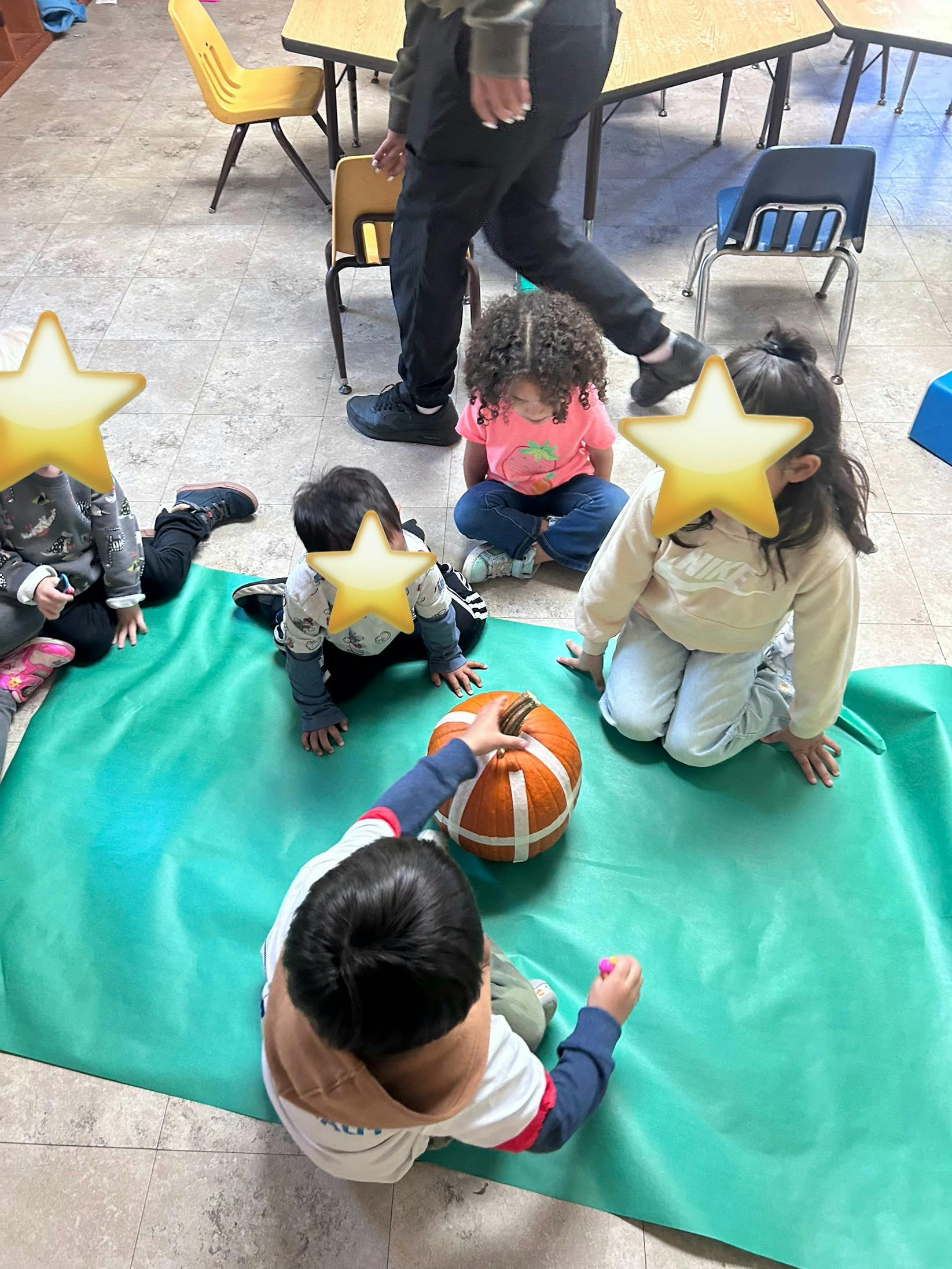 children on a green mat playing with a pumpkin, with a person standing nearby