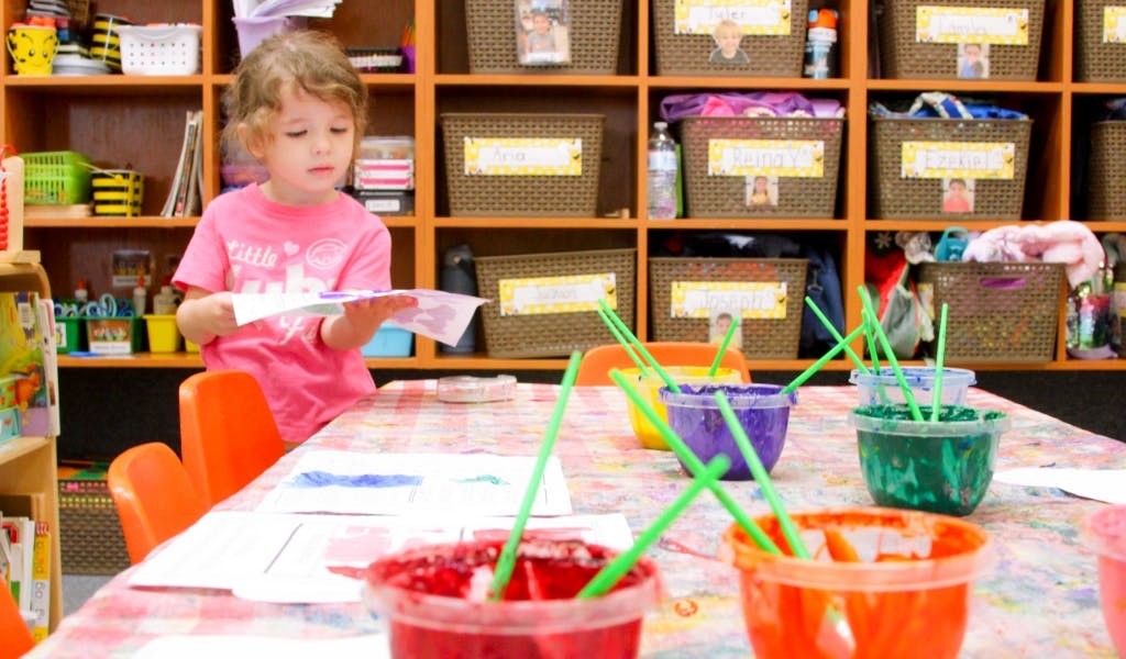 Child holding paper at a table with paint pots and brushes in a classroom