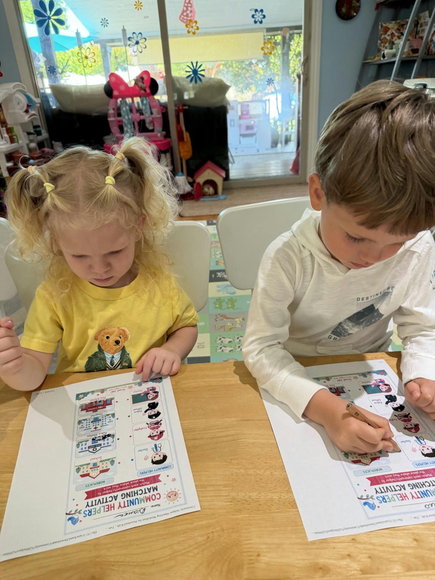 Two children working on community helper matching activity worksheets at a table