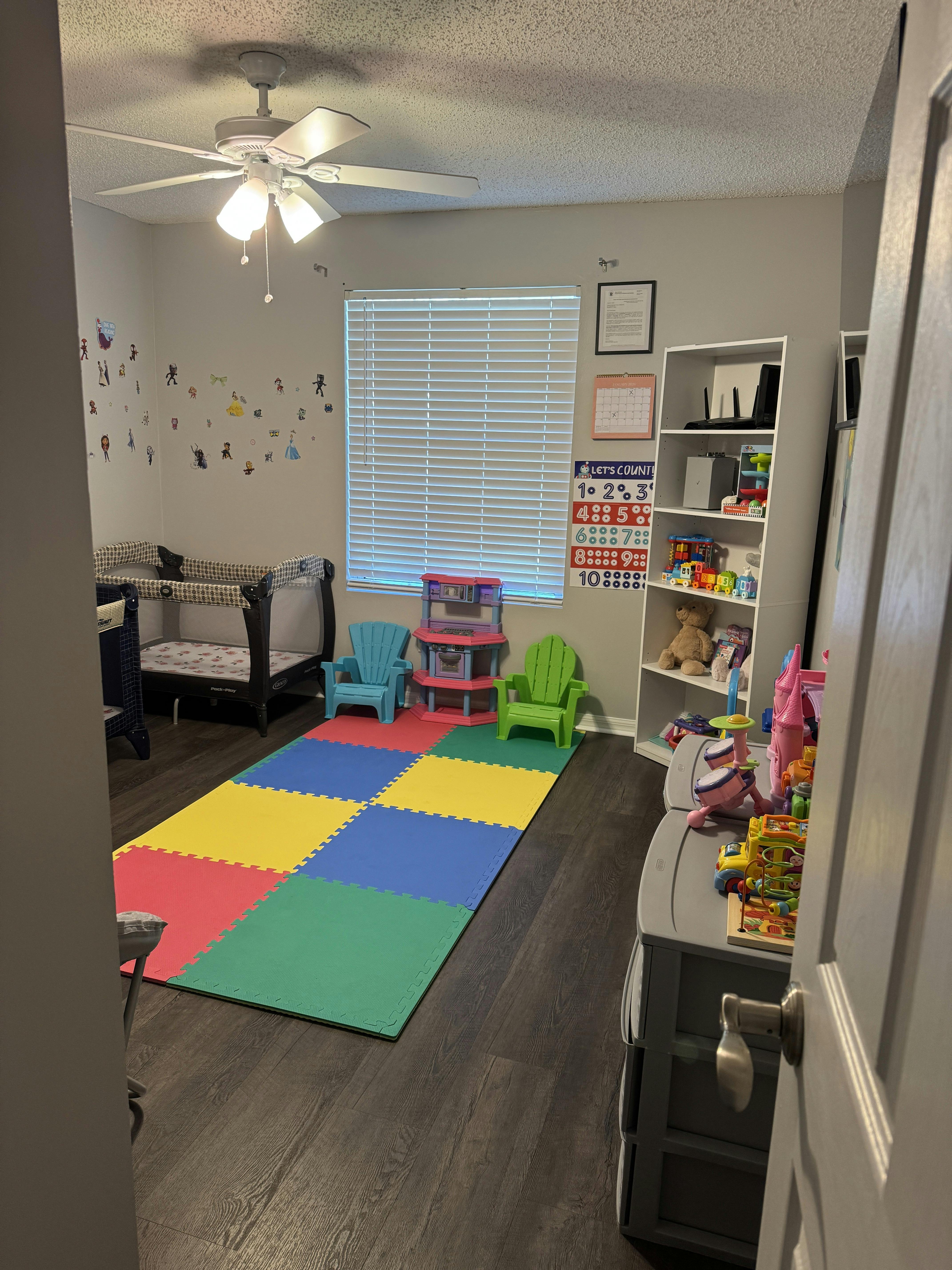 Room with cribs, colorful play mat, small chairs, and shelves with shelves of toys