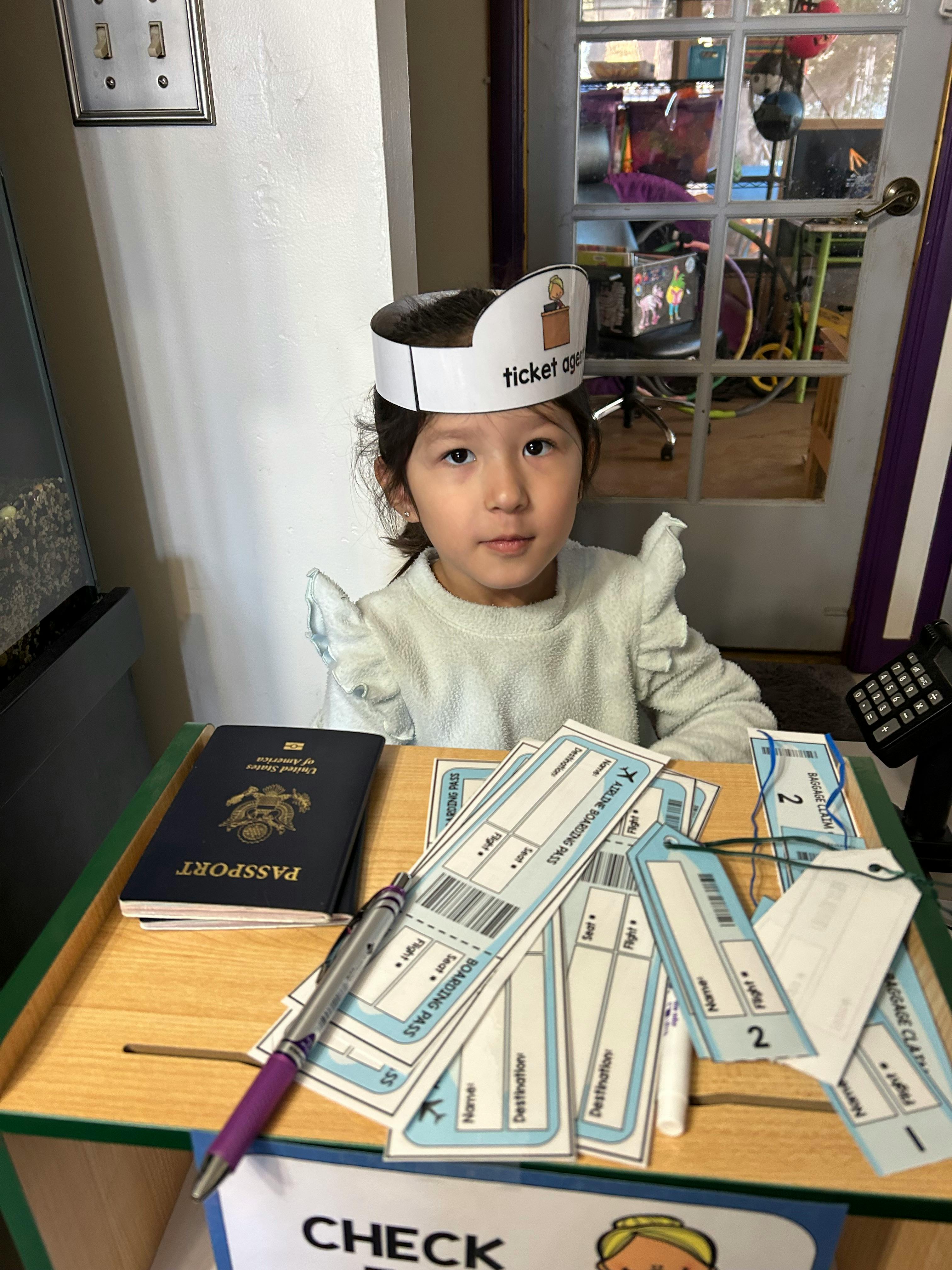Child wearing a paper hat with "ticket agent" text, with toy passport and tickets