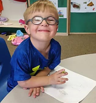 Child in glasses sitting at a table with paper and a writing utensil