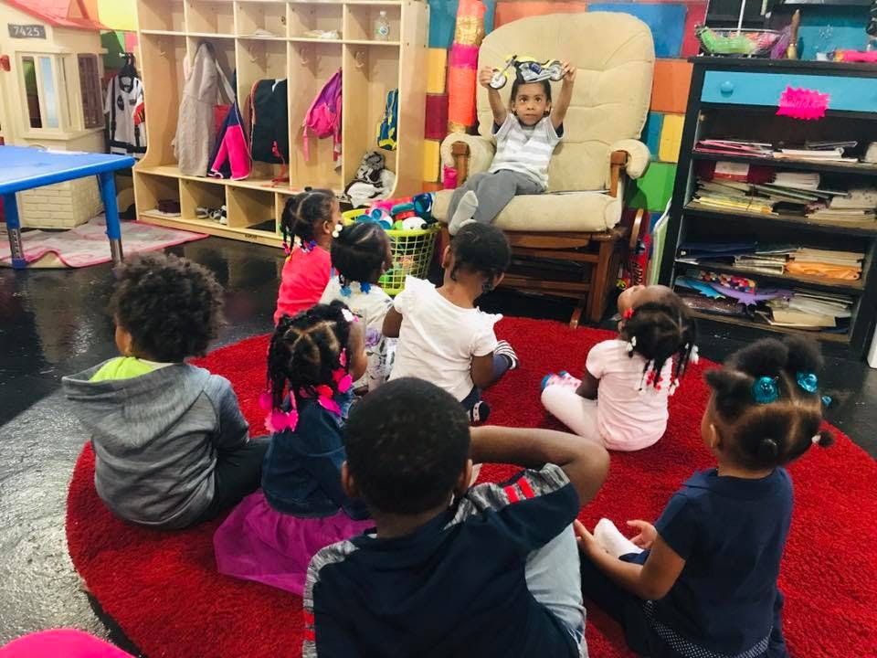 Children sitting on a red rug looking at a child in a chair holding an object