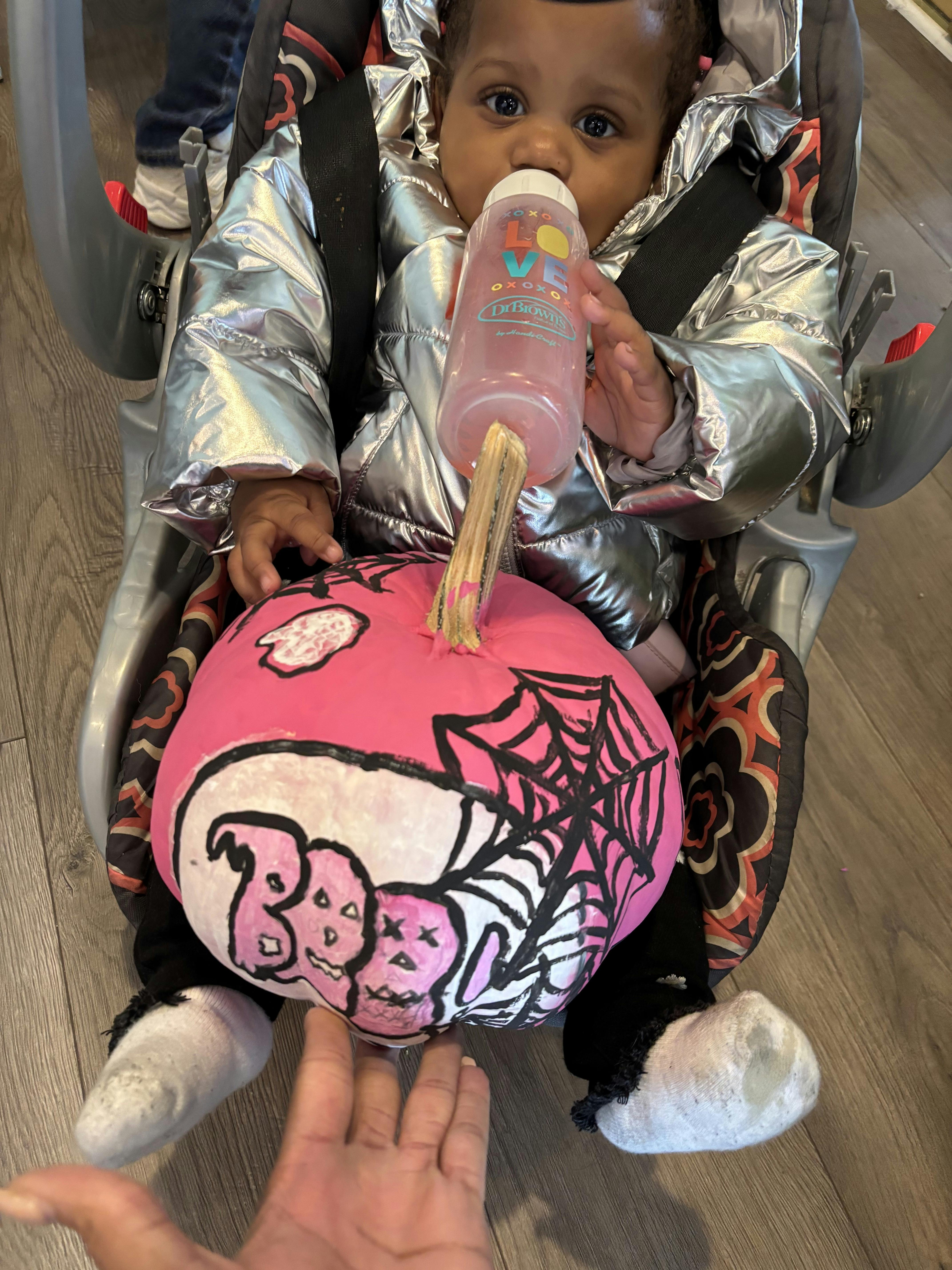 Child in a silver jacket drinking from a bottle, with a pink decorated pumpkin