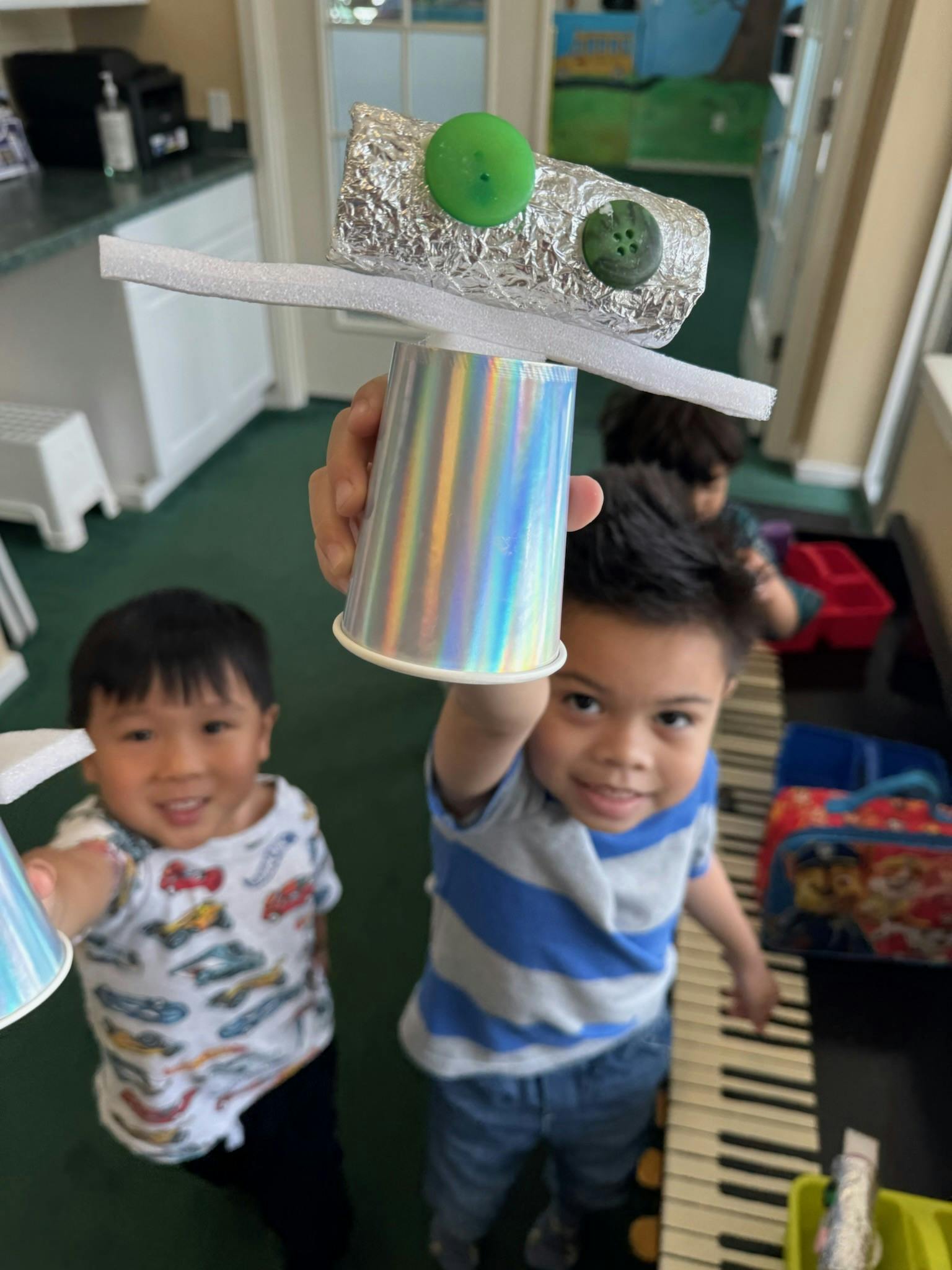 Child holding up a craft made from a cup and foil, another child smiling