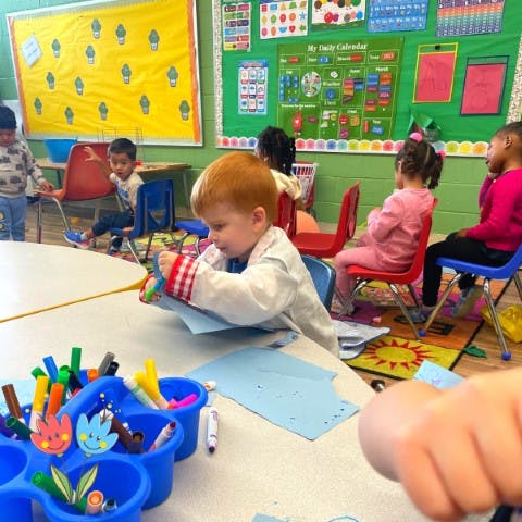 Child cutting paper at a table with other children in a classroom