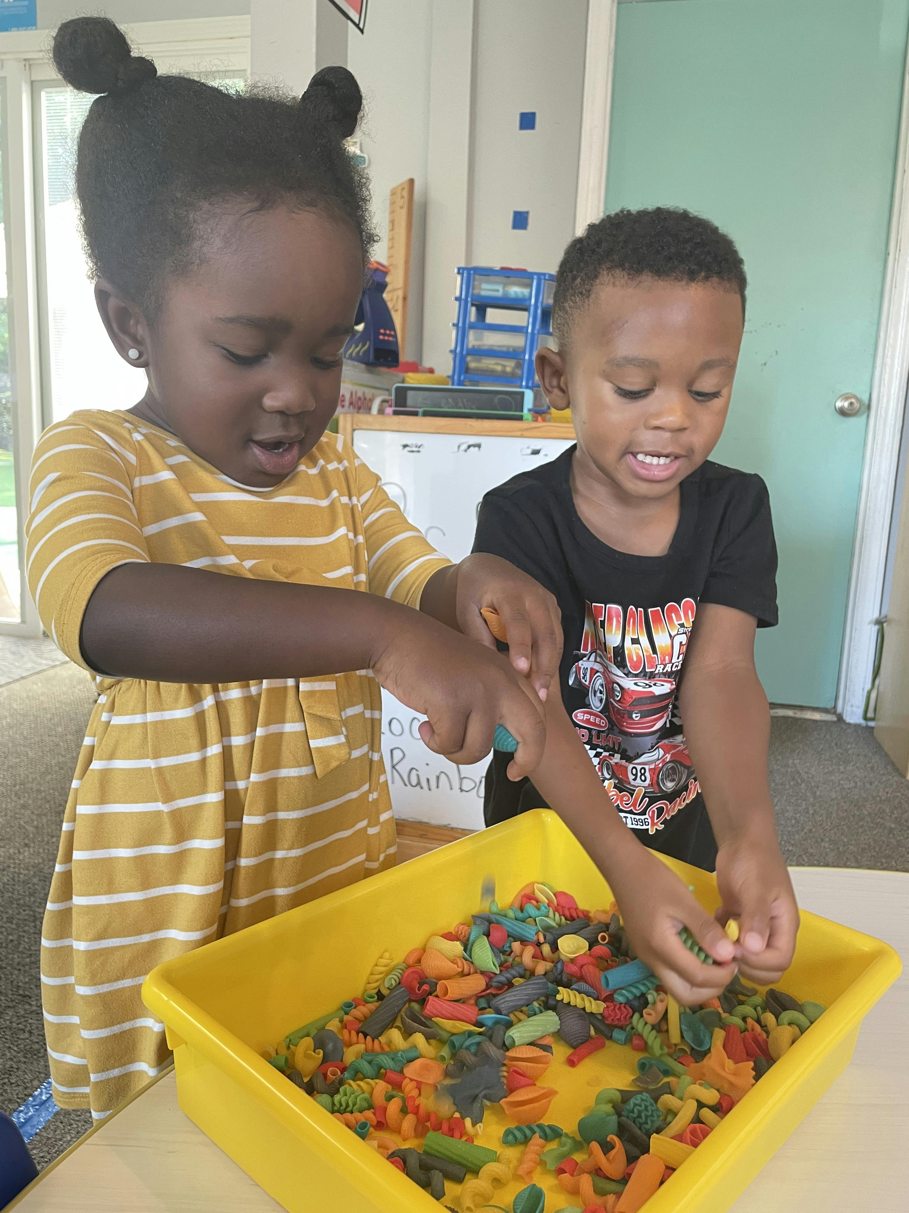 Two children playing with colorful pasta in a plastic bin