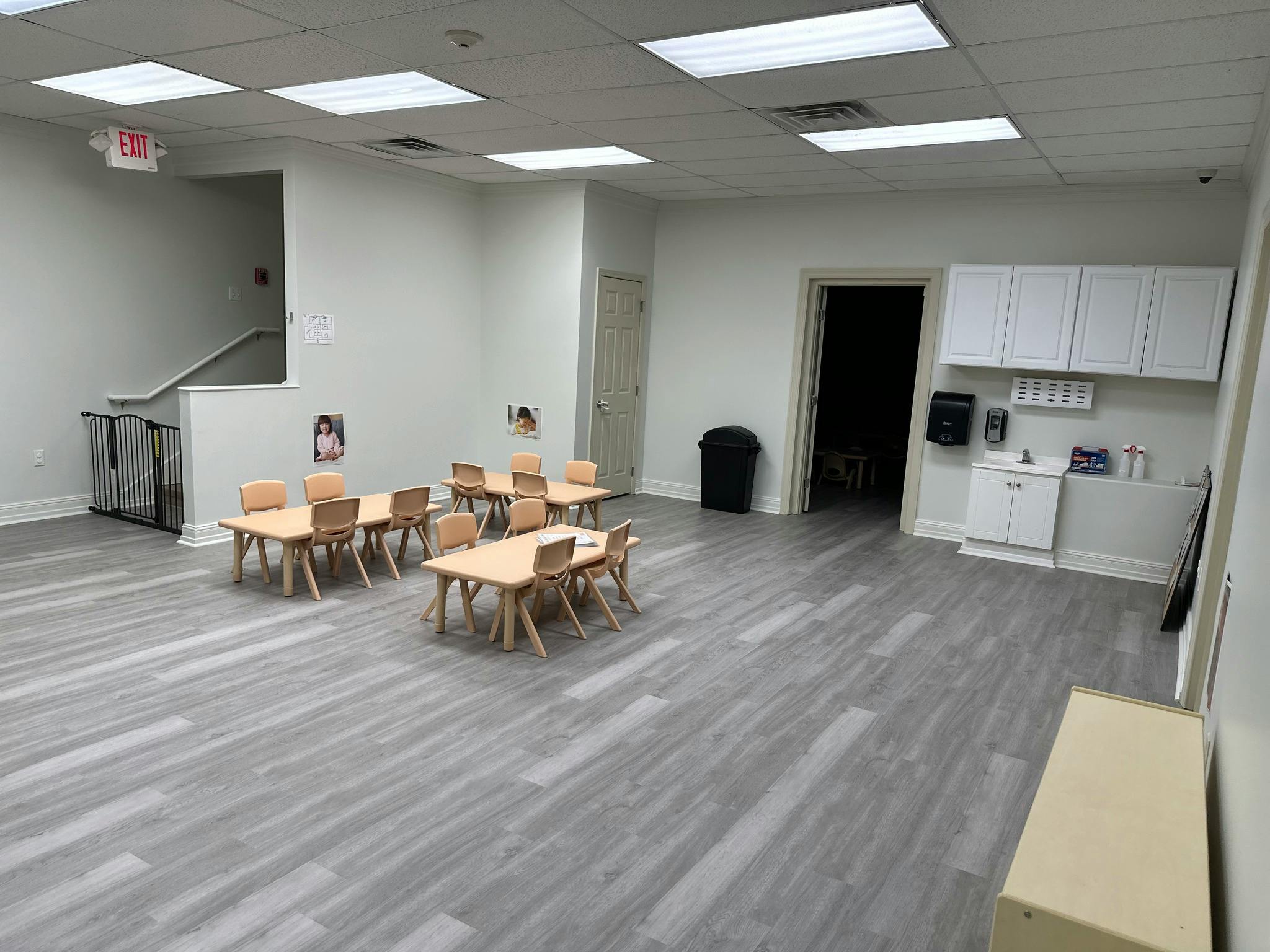 Empty classroom with small tables and chairs, a sink area, and a staircase