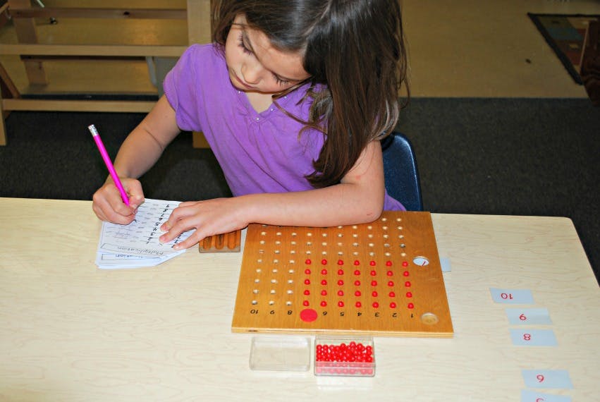 Child writing on paper with a pencil next to a multiplication board