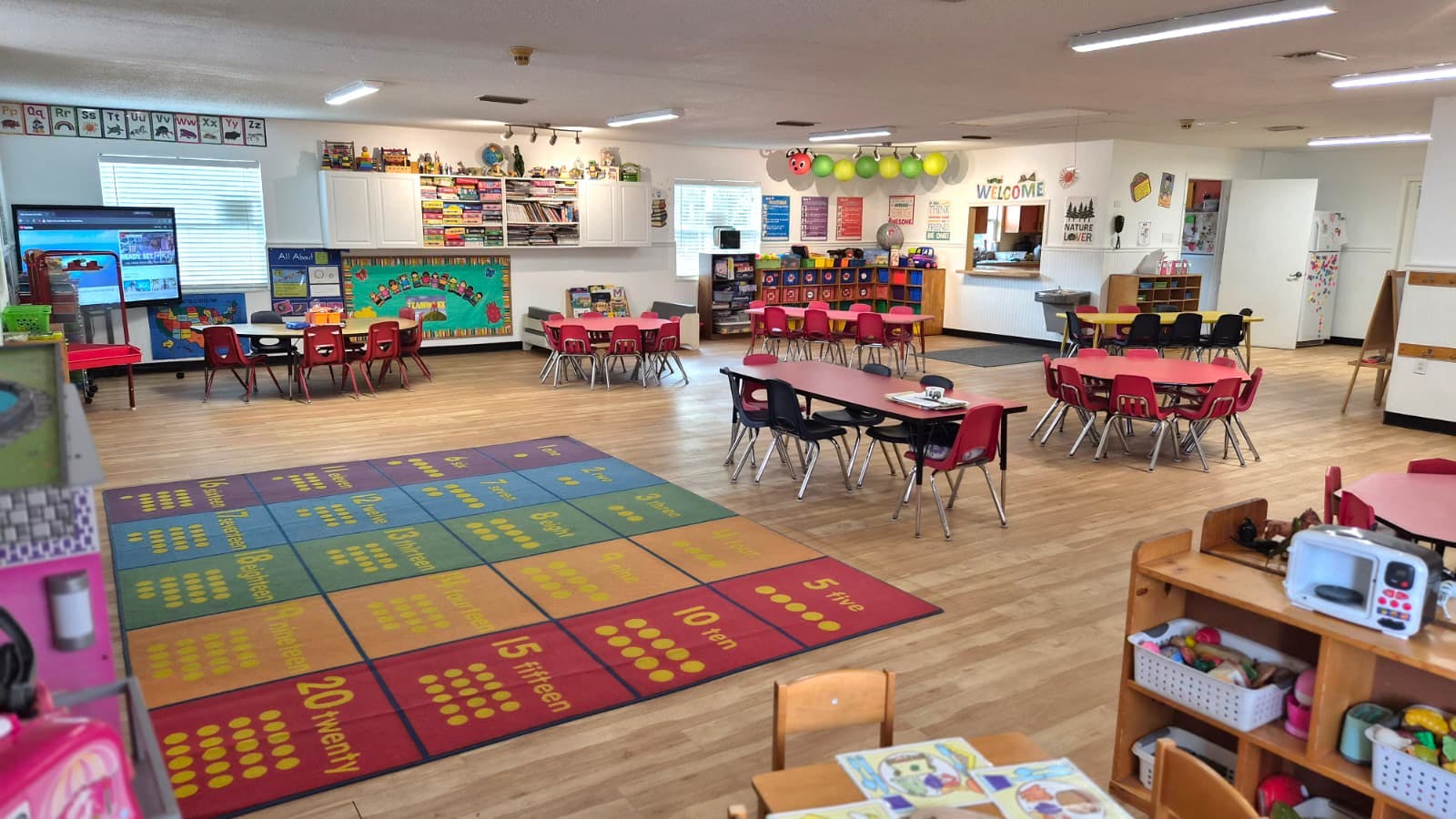 A colorful classroom with small tables and chairs, a large number rug, and learning materials