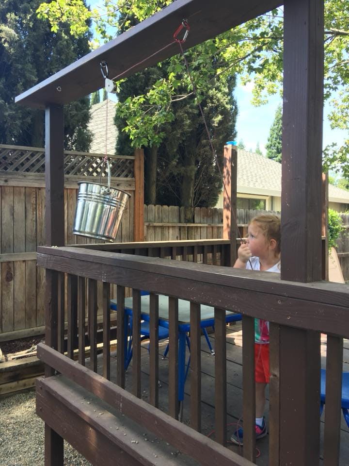 Child on a wooden play structure with a metal bucket hanging from a pulley