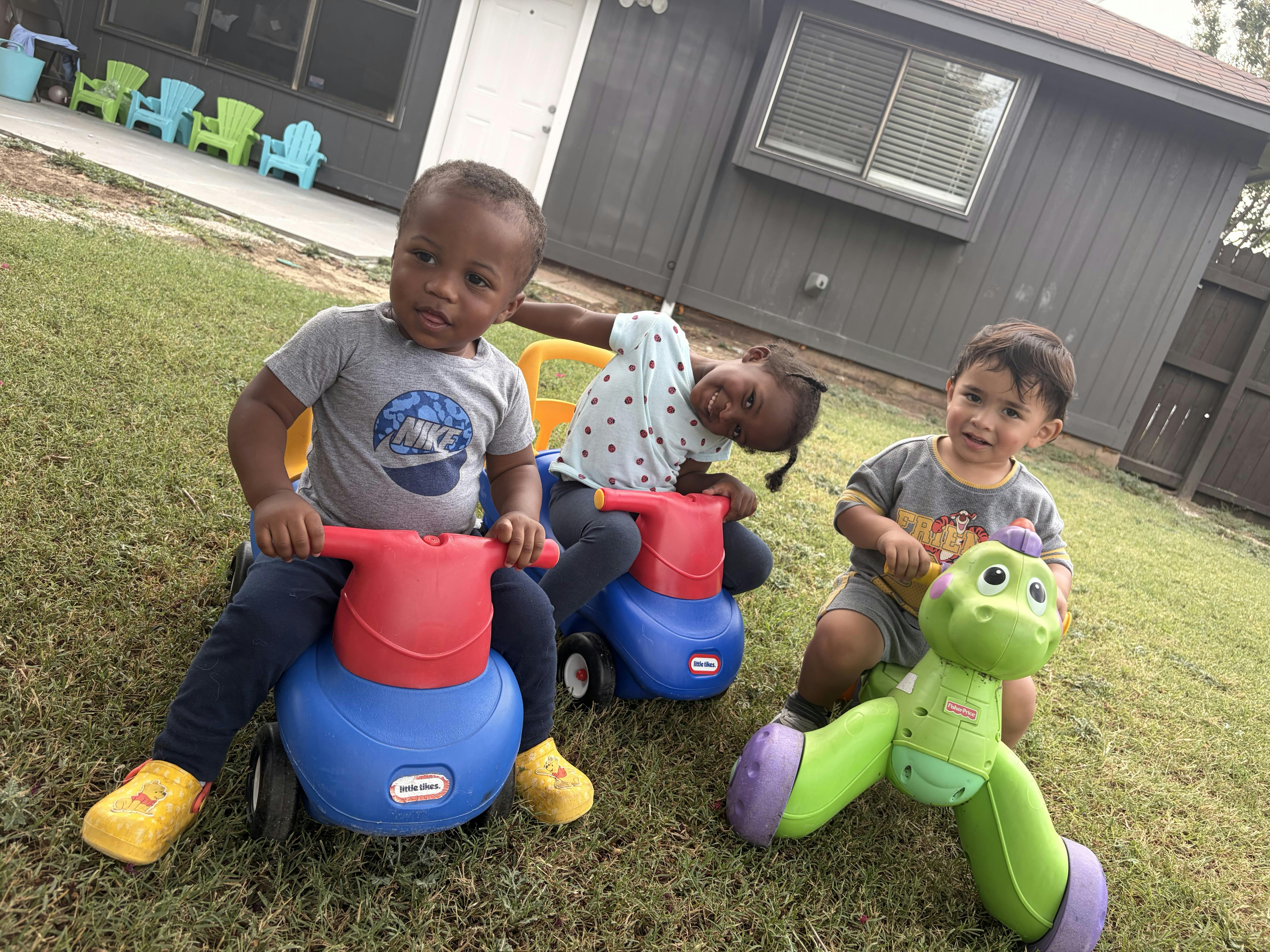 Three children riding on plastic toy vehicles on a lawn