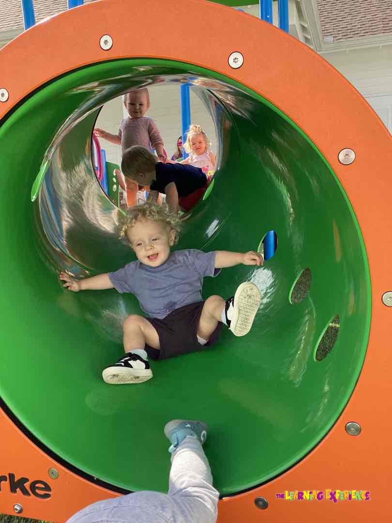 Child sliding down a green playground tube, other children visible in background
