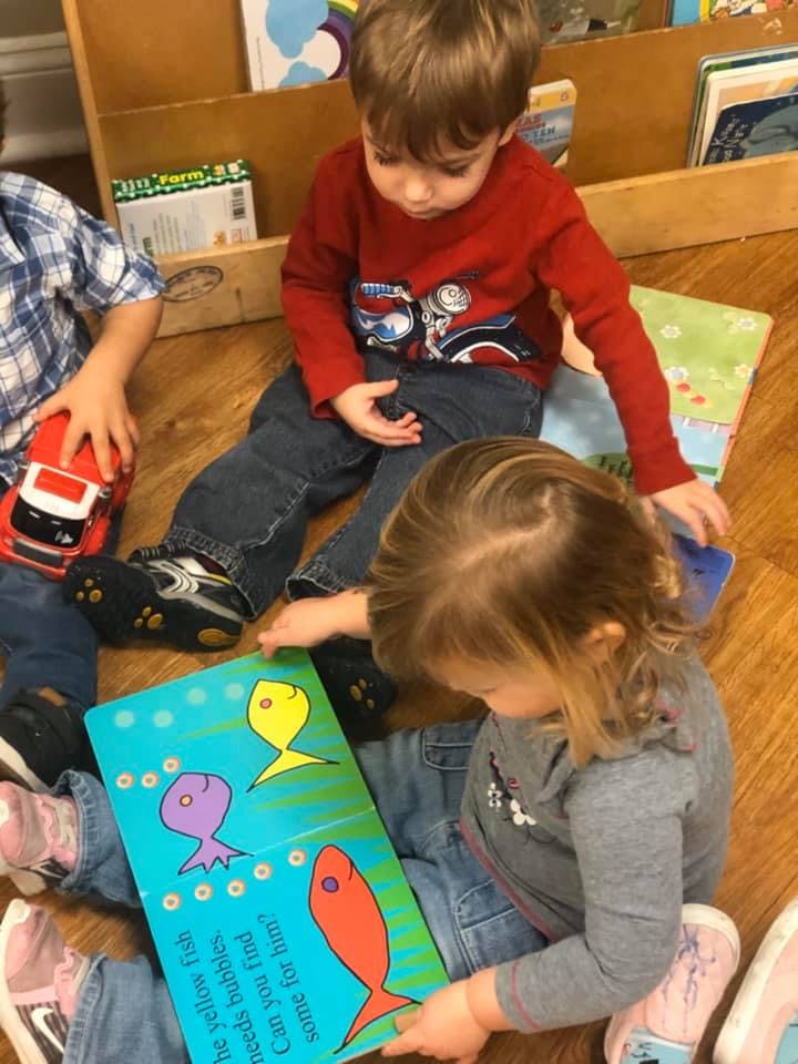 Three children sitting on the floor, one reading a book with fish illustrations