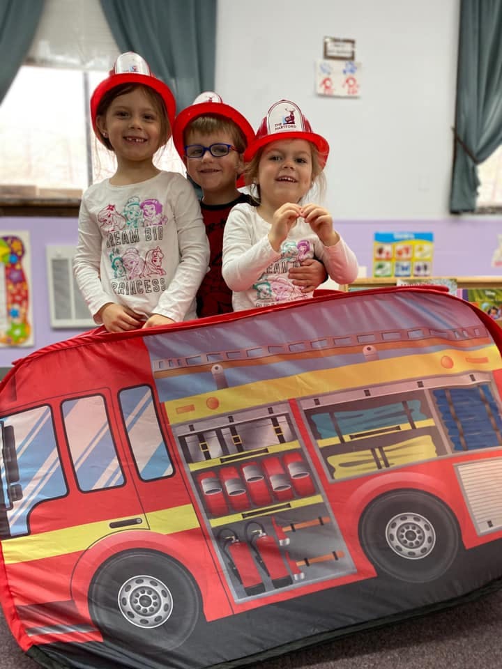 Three children wearing firefighter hats behind a toy fire truck