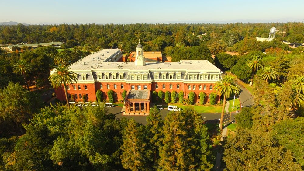 Large brick building with a white roof and cupola surrounded by trees