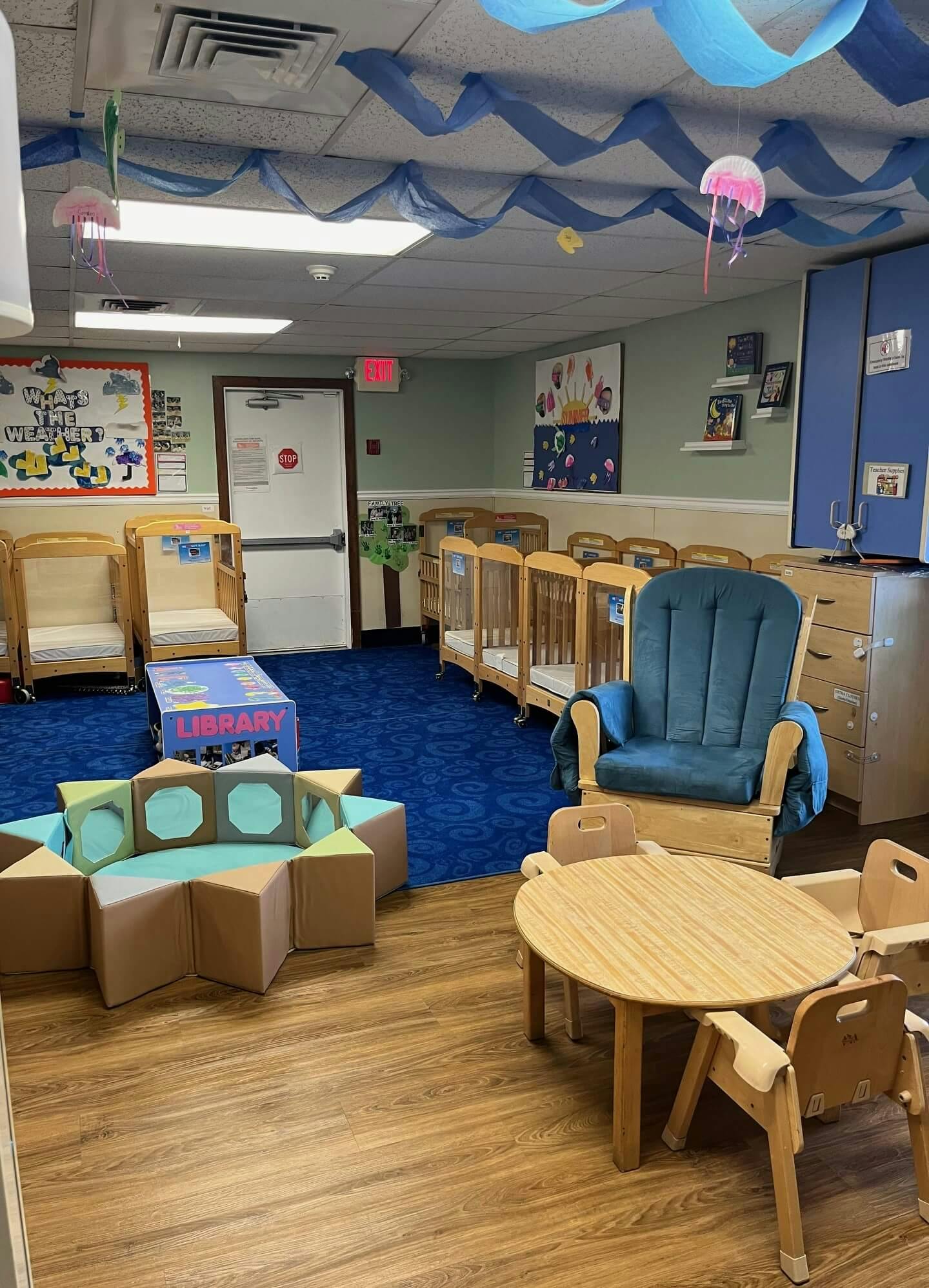 A classroom with cribs, a reading nook with text "LIBRARY", a rocking chair, and a small table with chairs