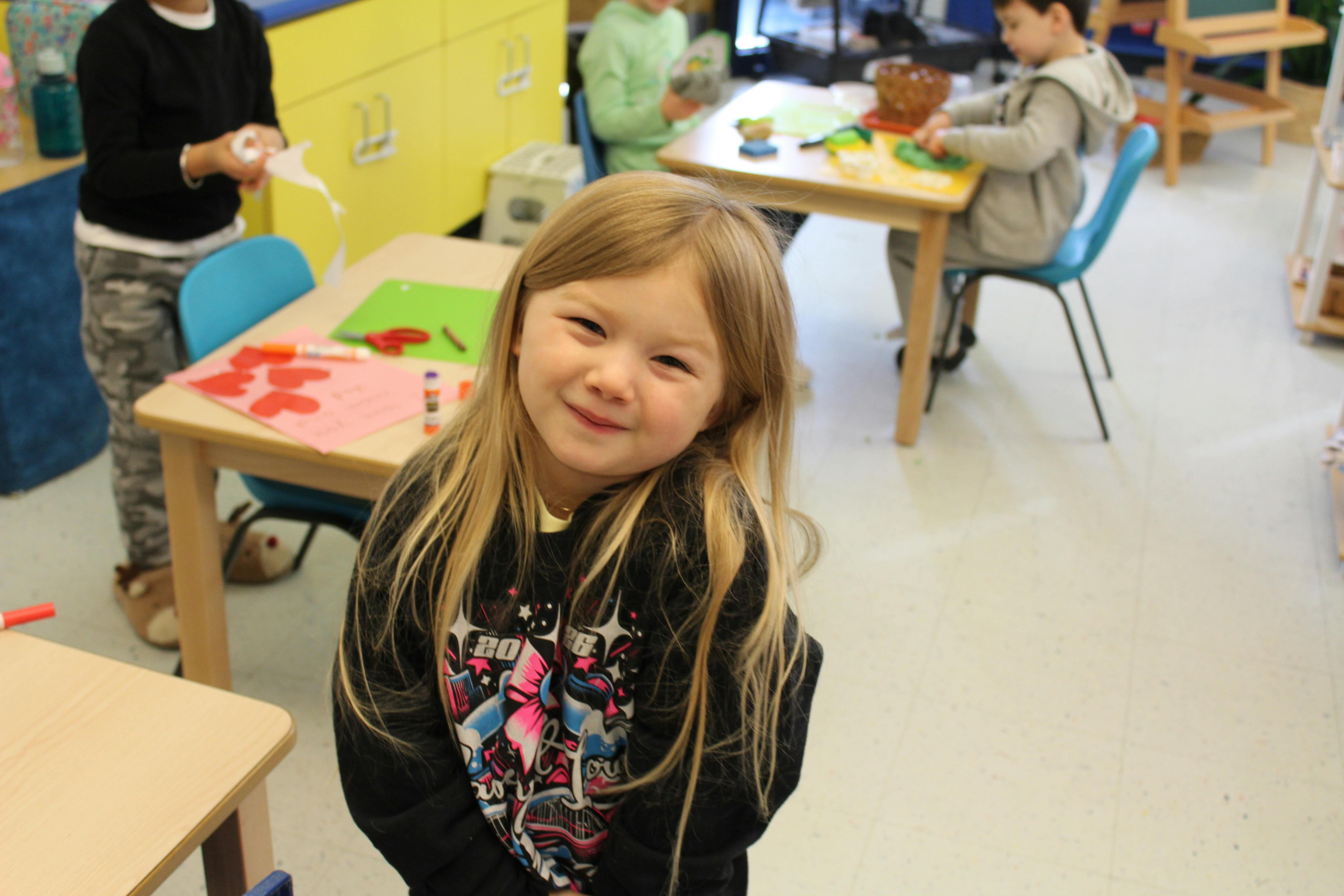 Child standing in a classroom with other children working at tables