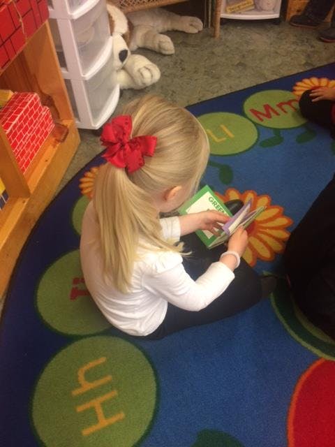 Child with blonde hair and red bow reading a book on a colorful rug