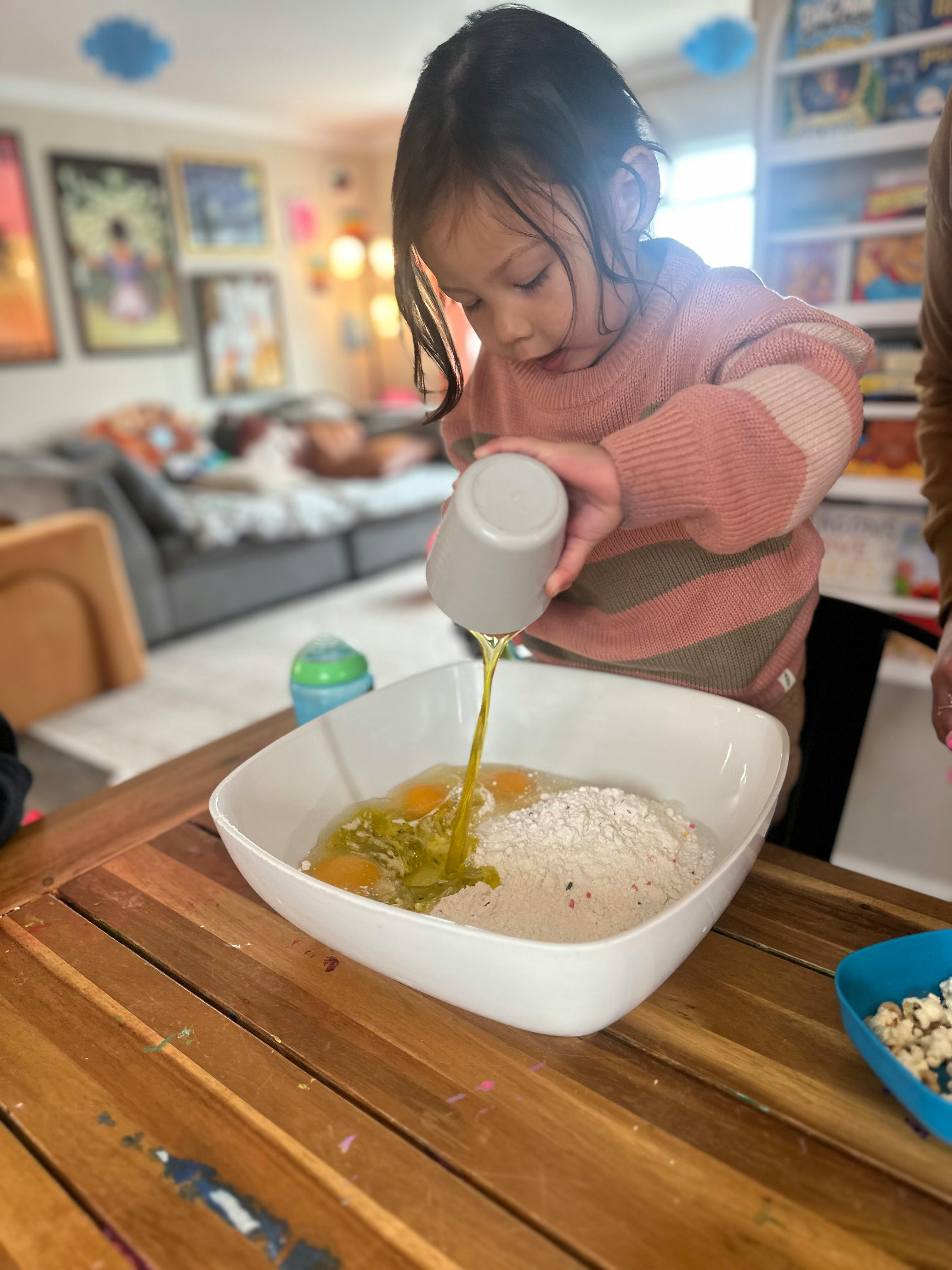 Child pouring liquid from a cup into a bowl with flour and eggs